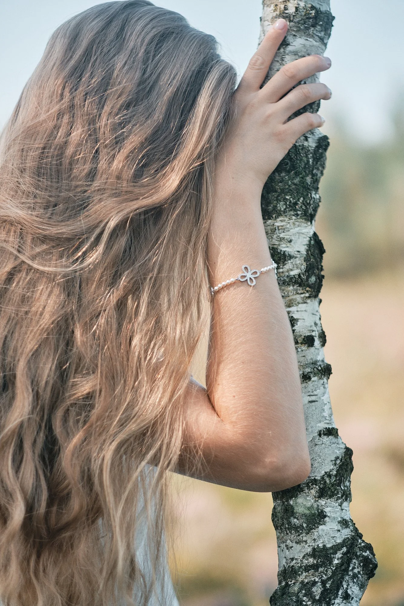 Eine Person mit langen, lockigen, hellbraunen Haaren, die eine Birke berührt, trägt ein silbernes Armband mit einer Blümchenform. Der Hintergrund ist unscharf und naturbelassen.