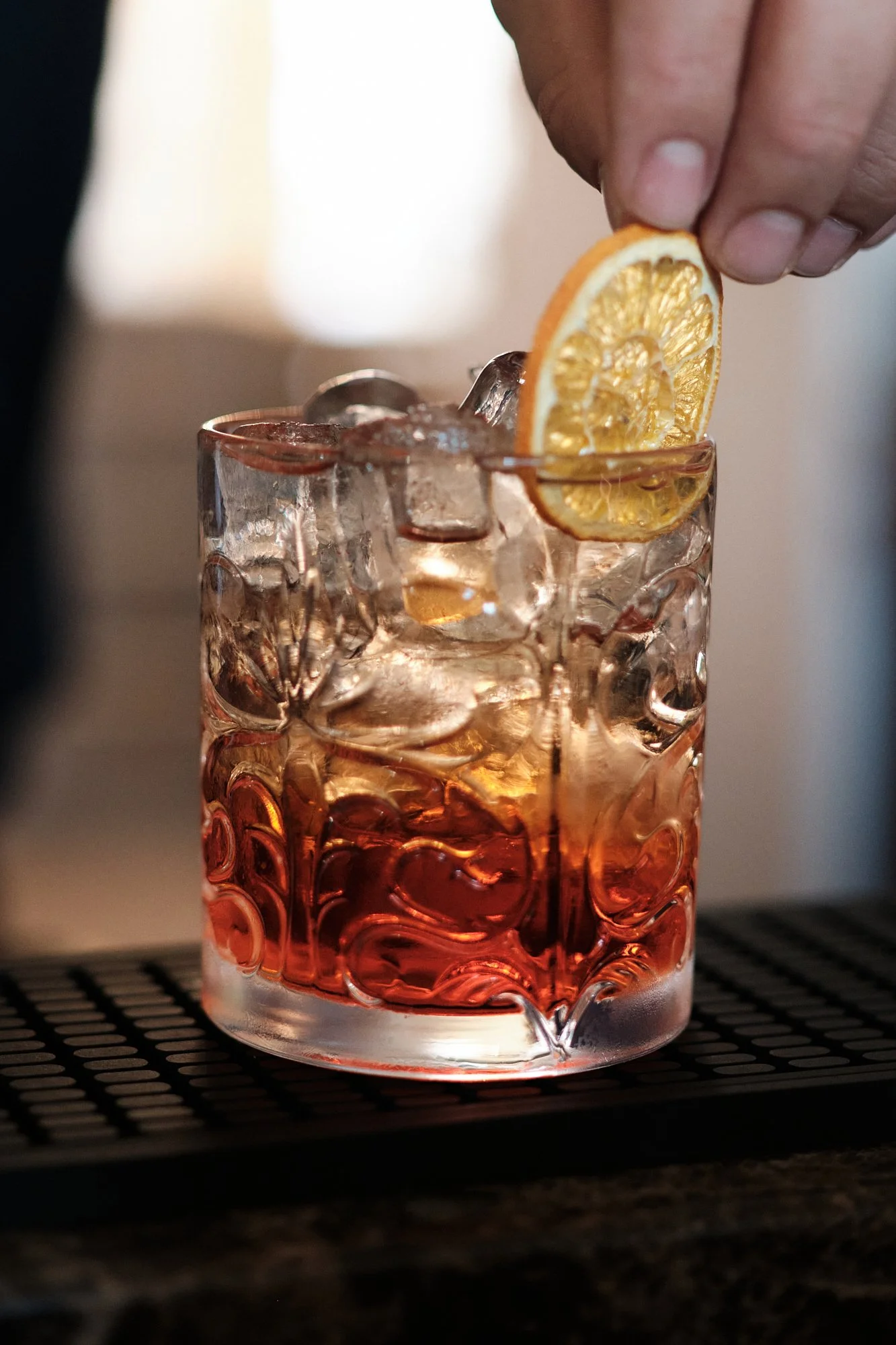 Ein Glas mit rotem Getränk und Eiswürfeln, eine Hand fügt eine Zitronenscheibe hinzu.