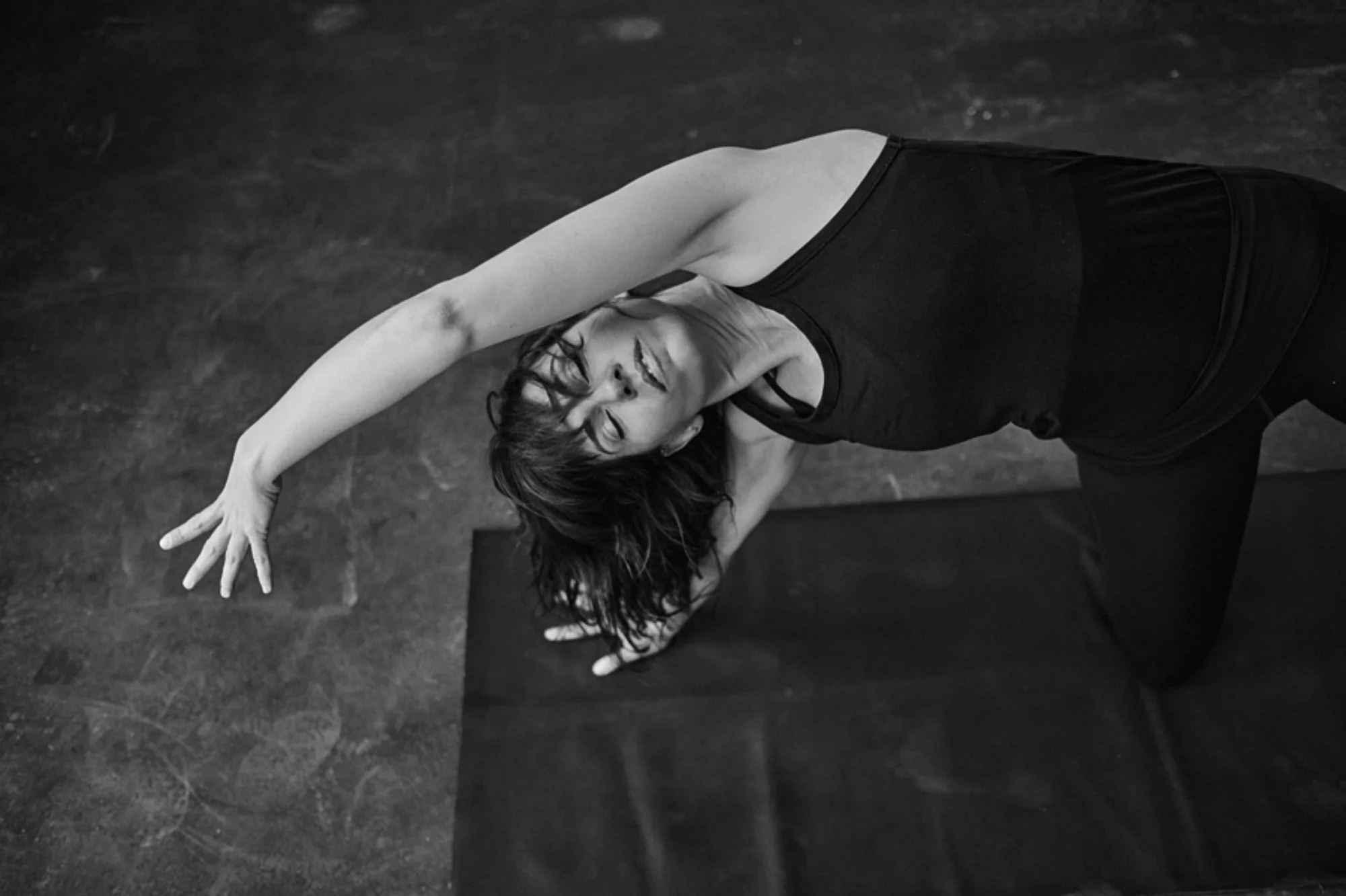 Frau in schwarzem Sportoutfit macht Yoga-Übung auf dunklem Boden, Birnenförmiger Körperhaltung, lächelnd, Augen geschlossen, Haare nach oben, entspannte Stimmung