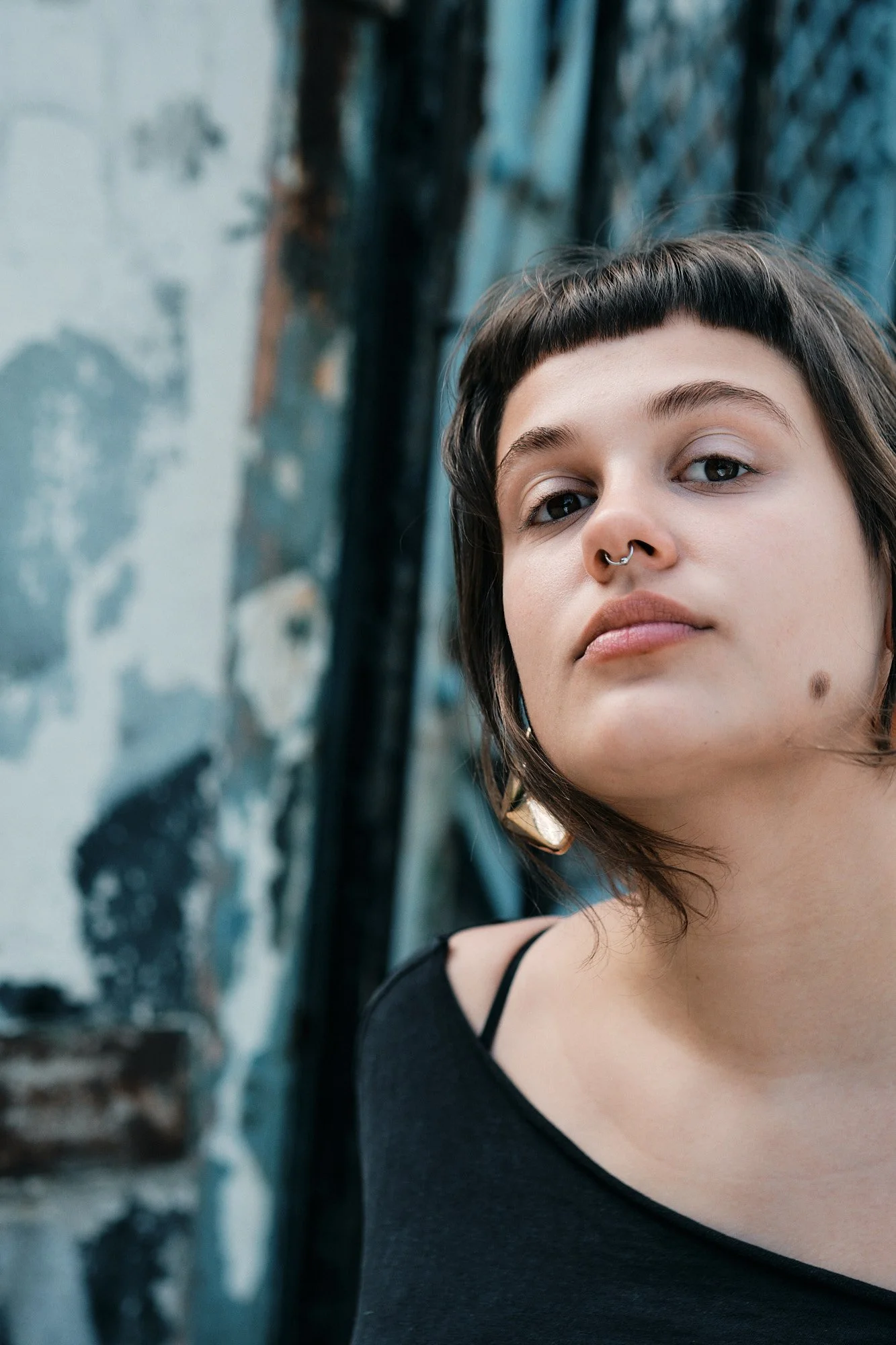 Ein junges Mädchen mit schwarzem Oberteil, kurzem Haar und Piercing an der Nase steht vor einer Wand mit Graffiti