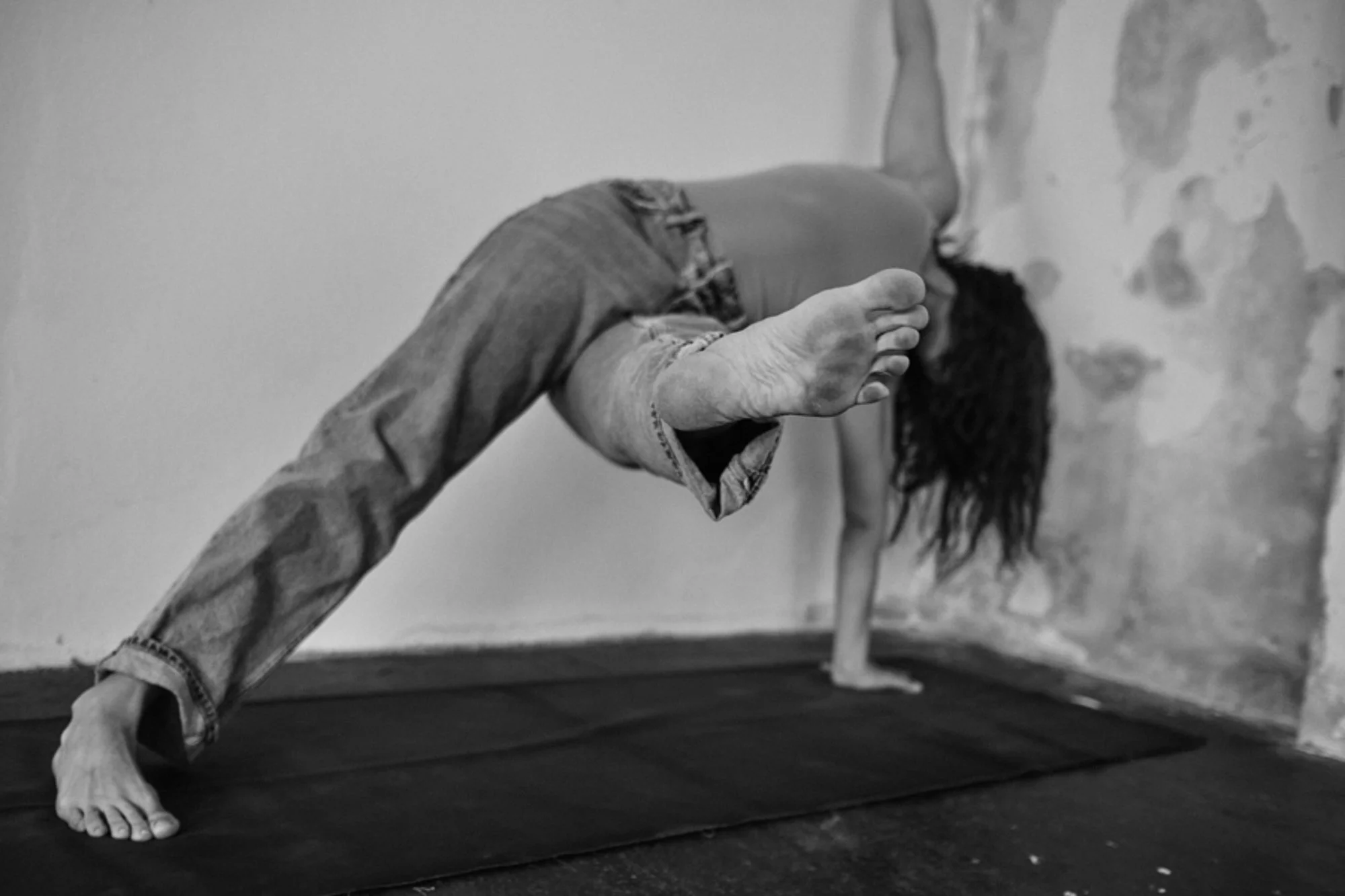 Schwarz-weiß-Fotografie einer nackten Person, die in einer Ballettpose steht, mit einem hochgestreckten Arm und einem Fuß auf einer Plattform, im Hintergrund eine Wand mit unregelmäßigen Farb- oder Putzflecken.