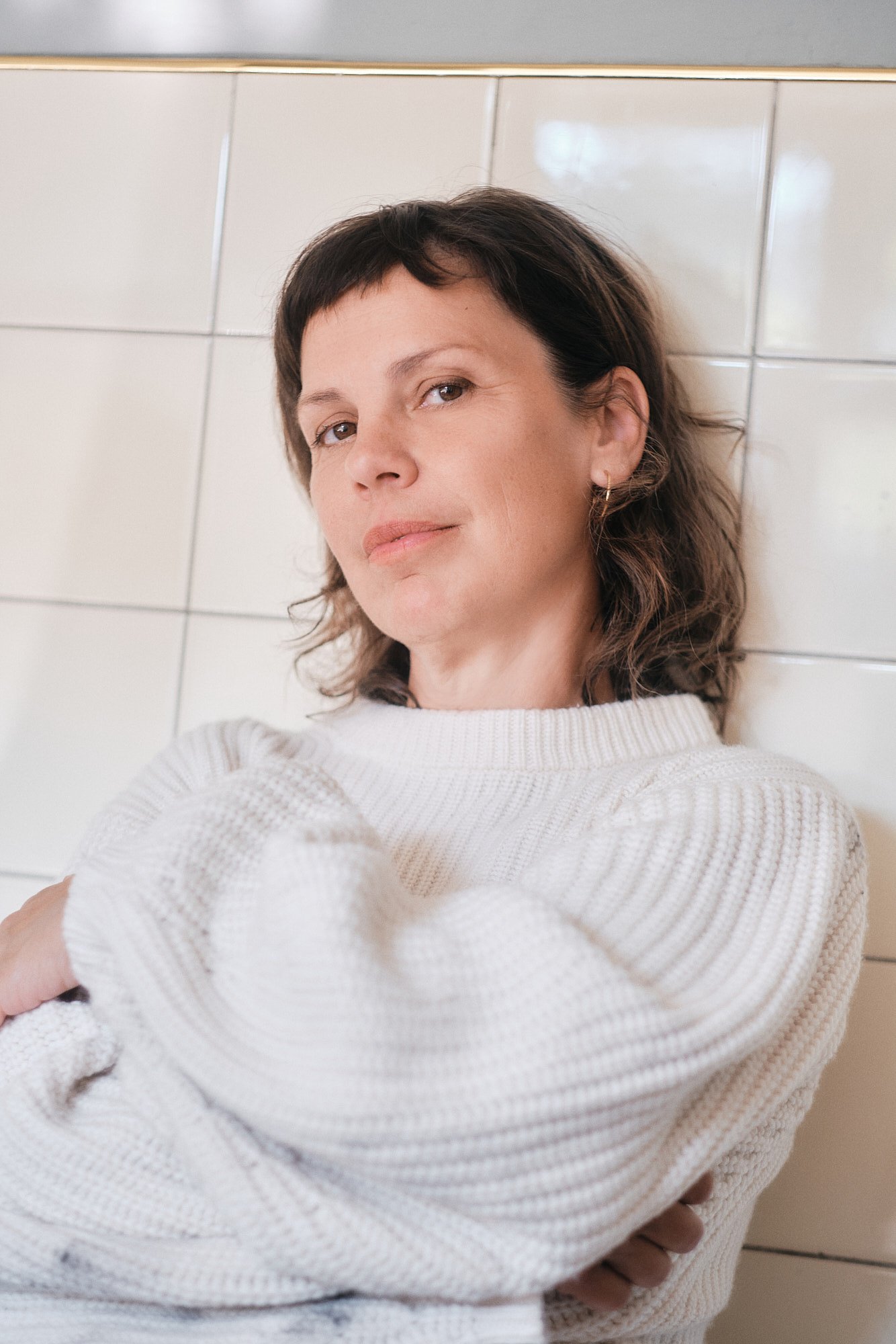 Frau mit brauen kurzen Haaren, trägt goldene Ohrringe und einen weißen Rippstrickpullover, lehnt an einer weißen Fliesenwand.