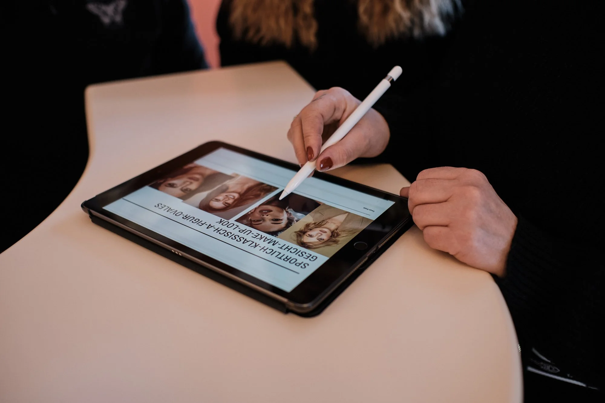 Eine Person benutzt einen digitalen Stift, um auf einem Tablet zu zeichnen. Das Tablet zeigt Bilder von Frauen. Der Tisch ist beige, im Hintergrund ist ein dunkler Raum.