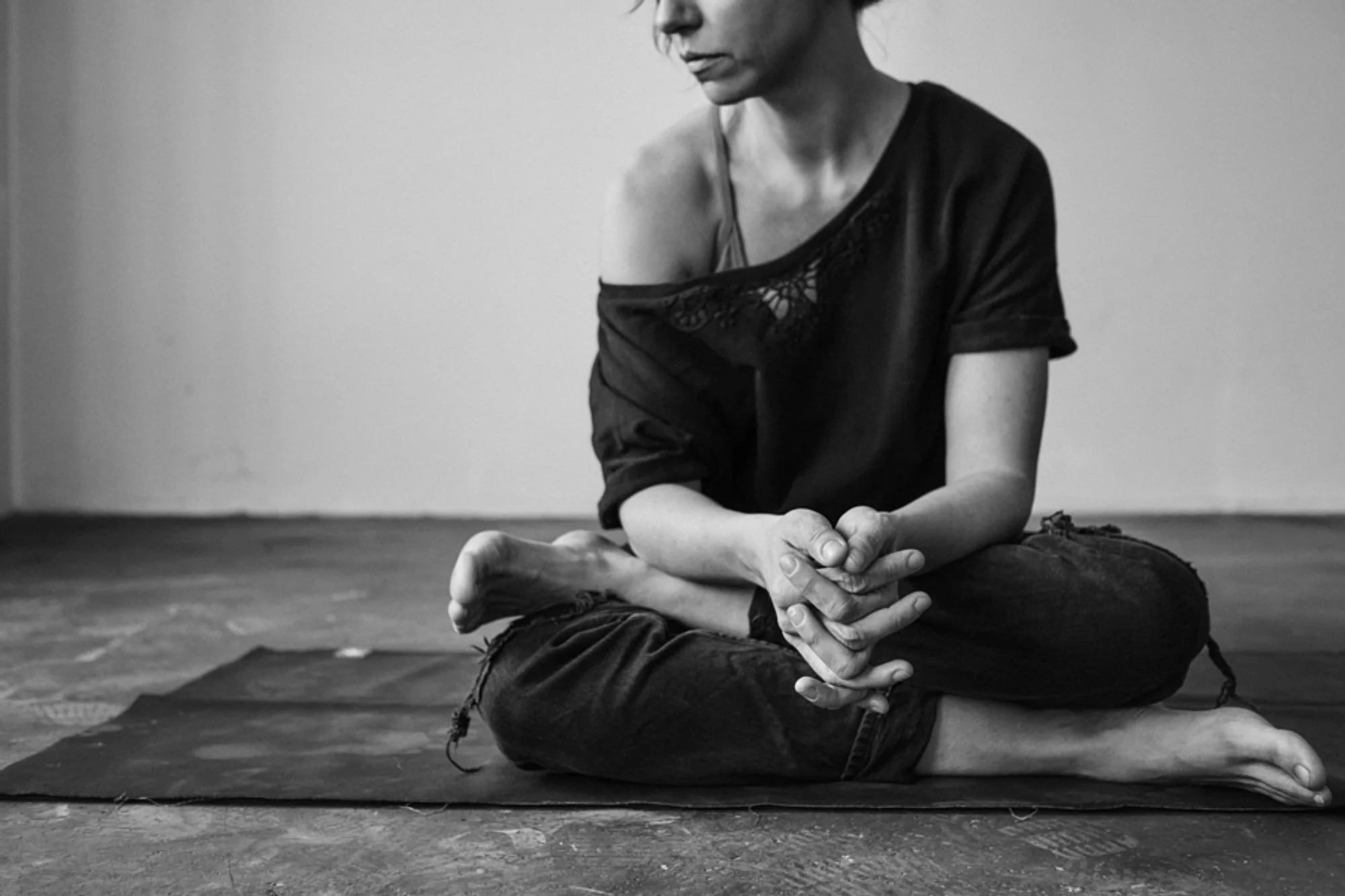 Schwarz-weiß Foto einer Frau beim Yoga, die in einer sitzenden Meditationshaltung auf einem Yoga-Teppich sitzt. Sie trägt ein lockeres Oberteil und legte ihre Hände in den Schoß, während sie nach vorne schaut.