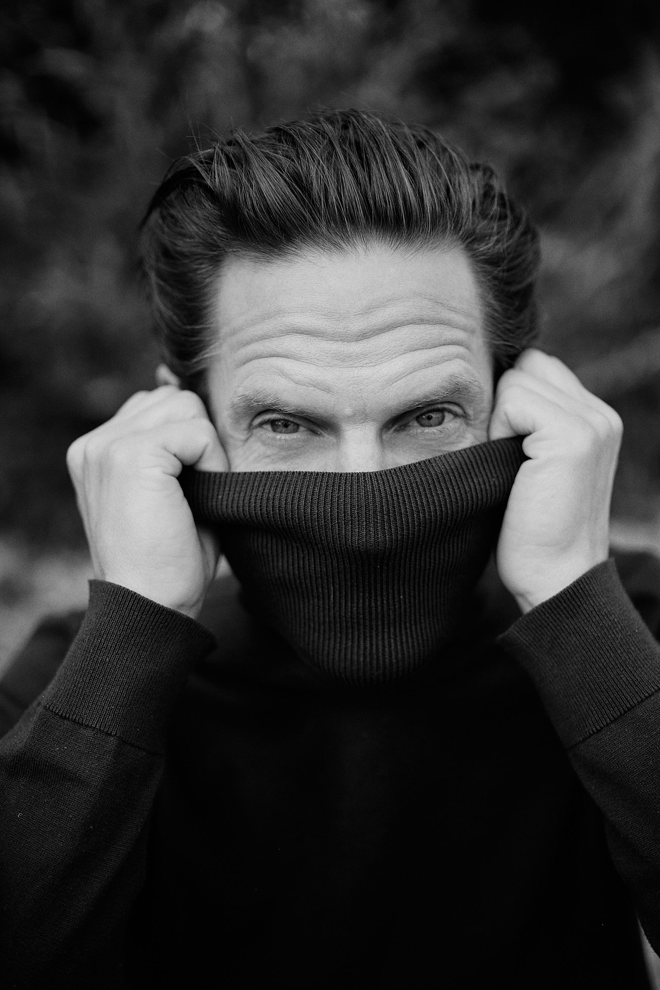 Schwarz-weiß-Foto eines Mannes, der einen schwarzen Rollkragenpullover trägt und die Kapuze hochzieht, sodass nur die Augen sichtbar sind, mit Blick direkt in die Kamera.