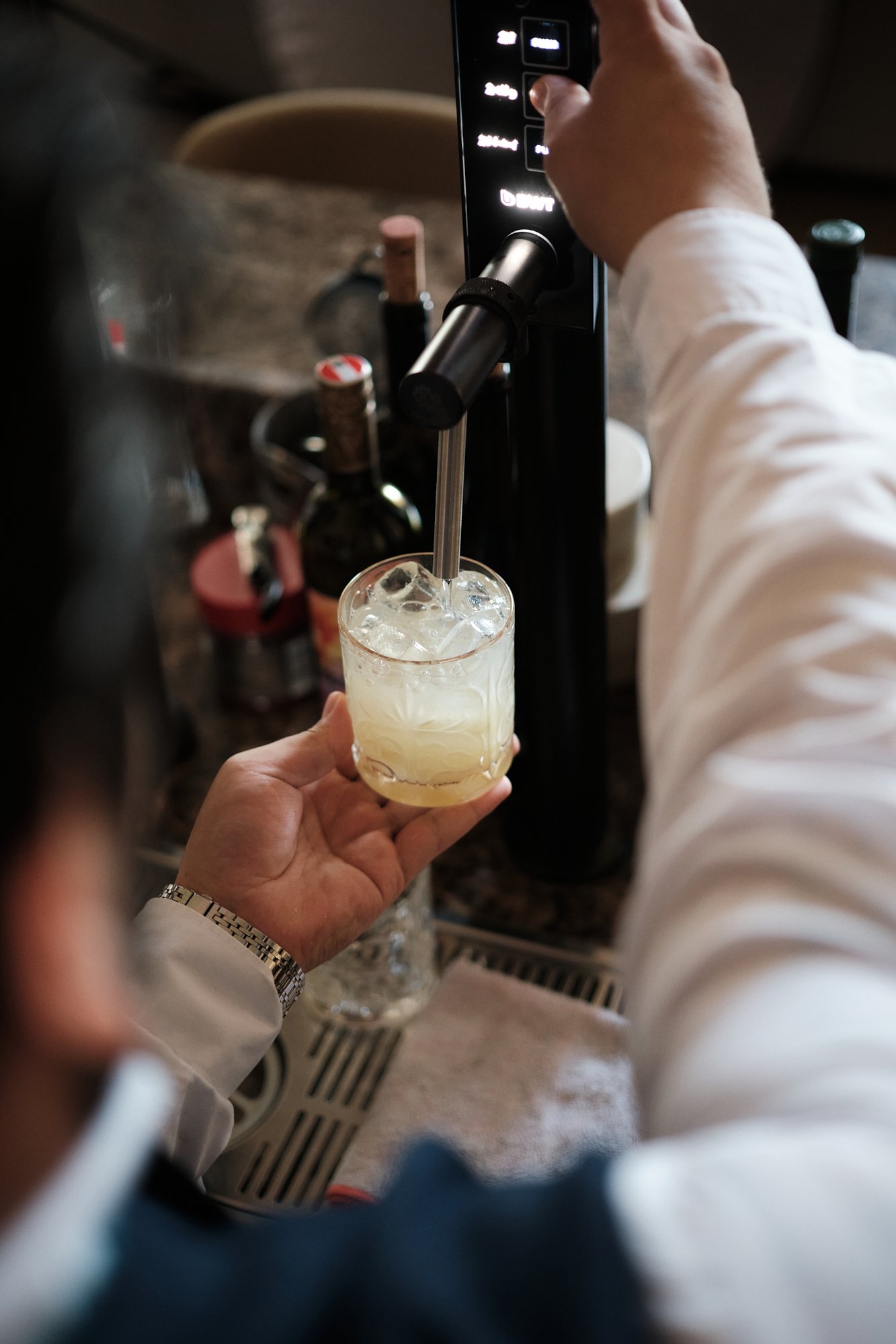 Eine Person gießt eine helle neumodische Trinkflüssigkeit in ein Glas mit Eis, wobei die Hand der Person sichtbar ist, die das Glas hält, während die andere Hand eine Maschine bedient.
