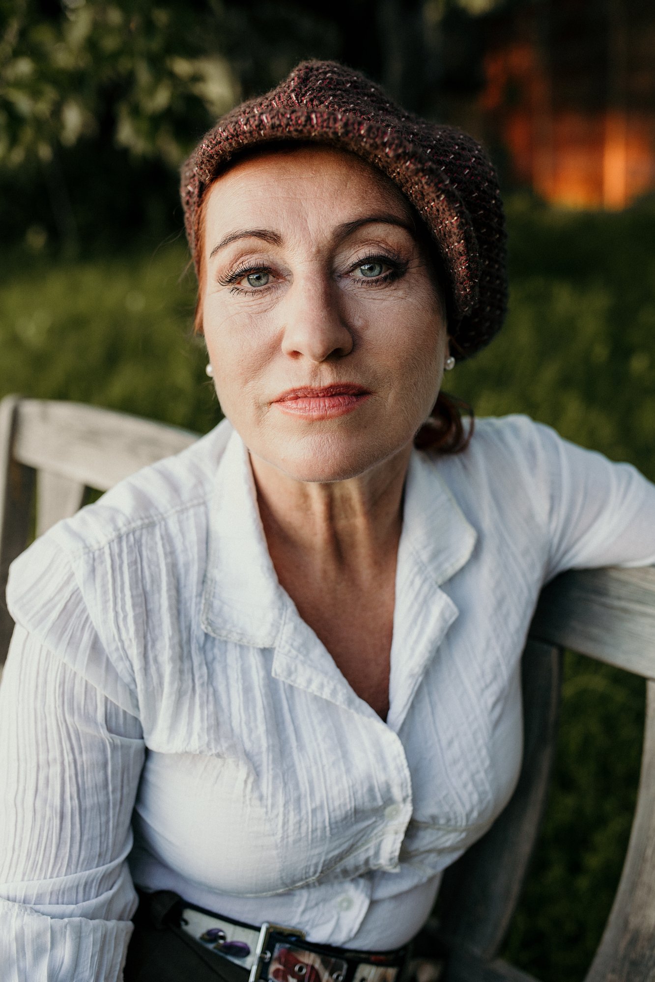 Nahaufnahme eines älteren Frauengesichts mit blauen Augen, rotem Haar, tragend eine braune Strickmütze, im Hintergrund ist grünes Laub bei Sonnenuntergang sichtbar.