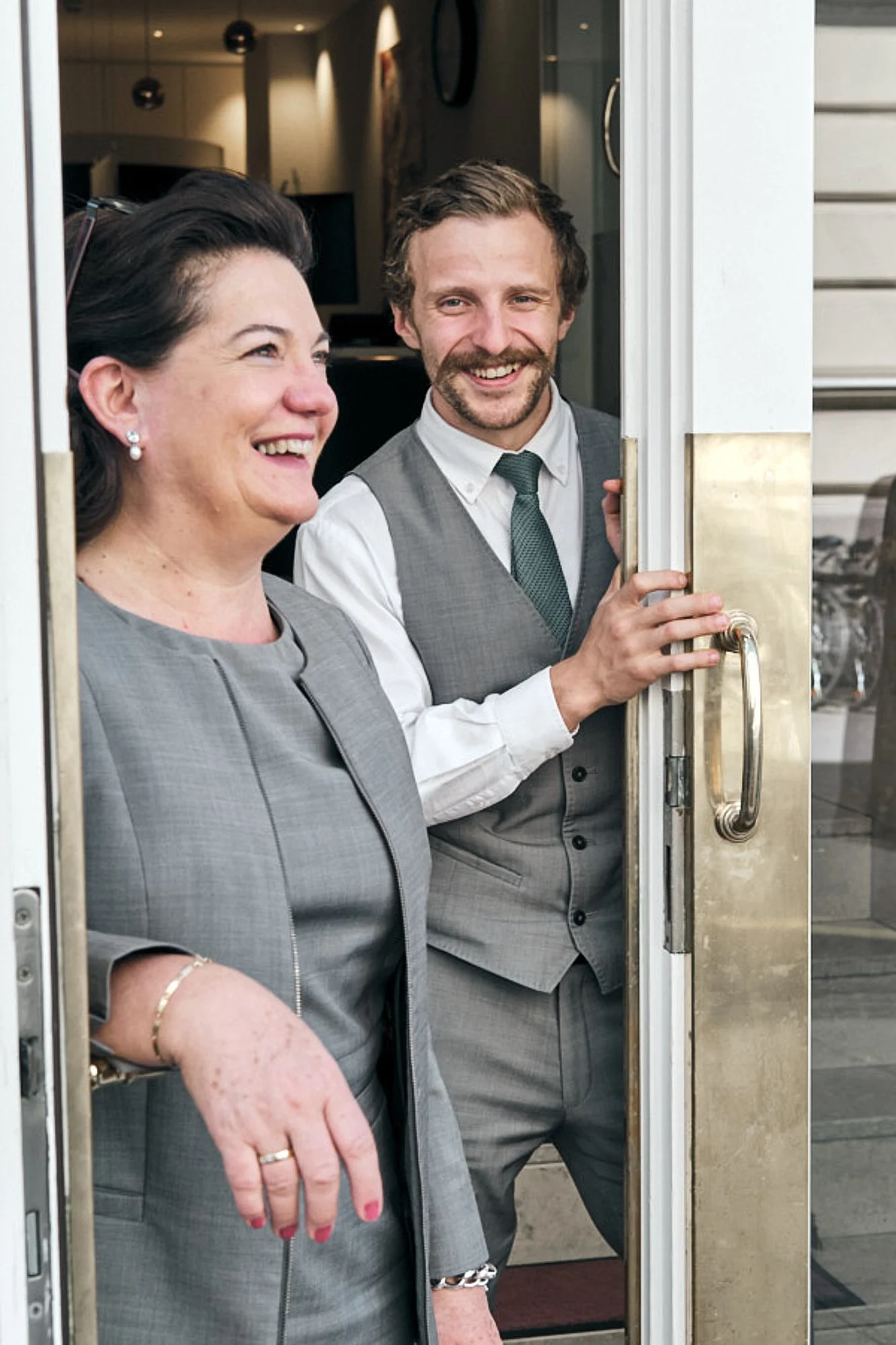 Zwei lachende Menschen, eine Frau und ein Mann, stehen an einem offenen Türrahmen, vermutlich bei einem Ereignis. Die Frau trägt einen grauen Anzug, die Hand ist sichtbar. Der Mann trägt einen grau-weißen Anzug mit schwarzer Krawatte und hält die Tür