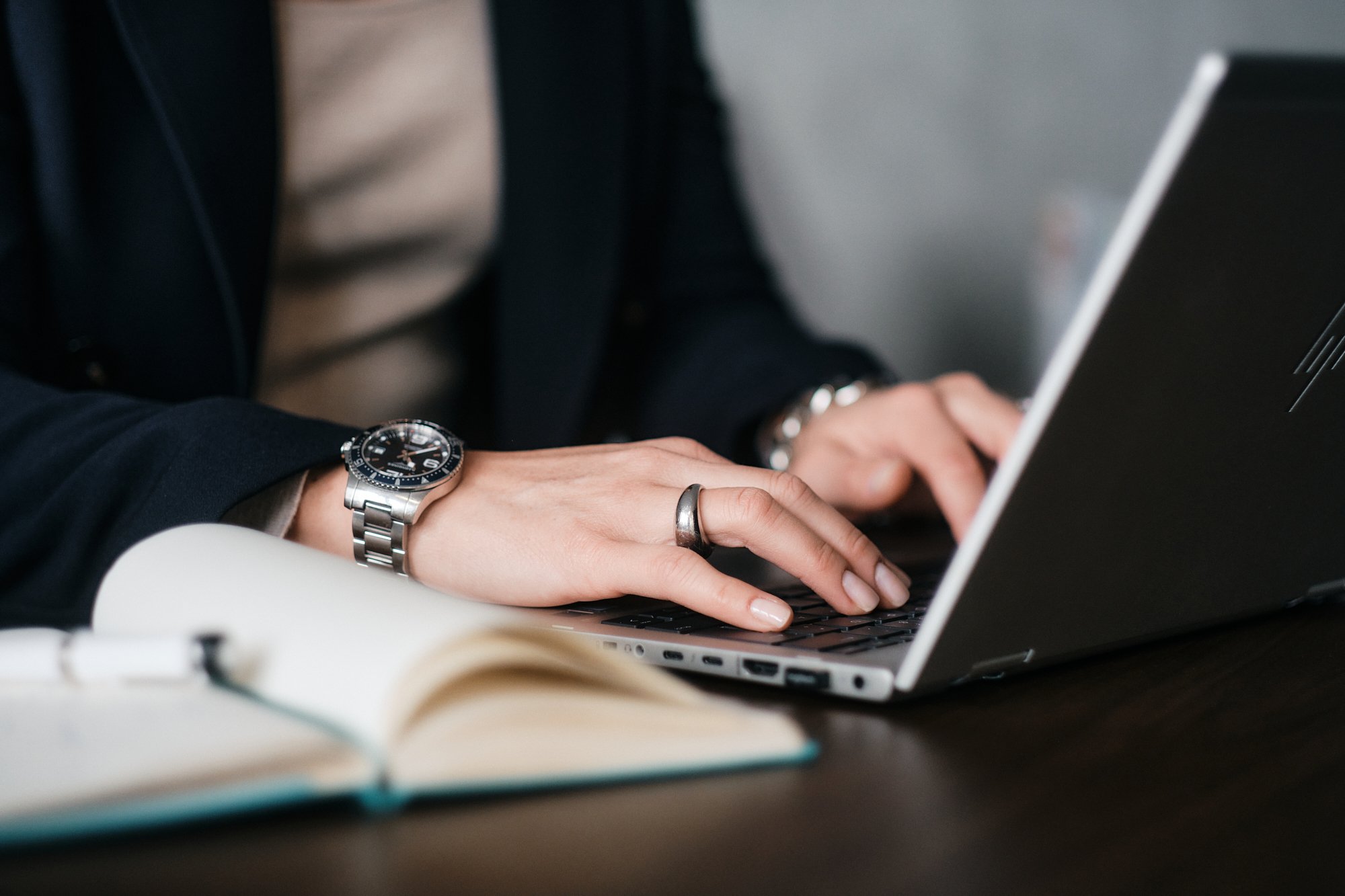 Person tippt auf einem Laptop, trägt eine Armbanduhr und einen Ring, im Hintergrund ein Notizbuch.