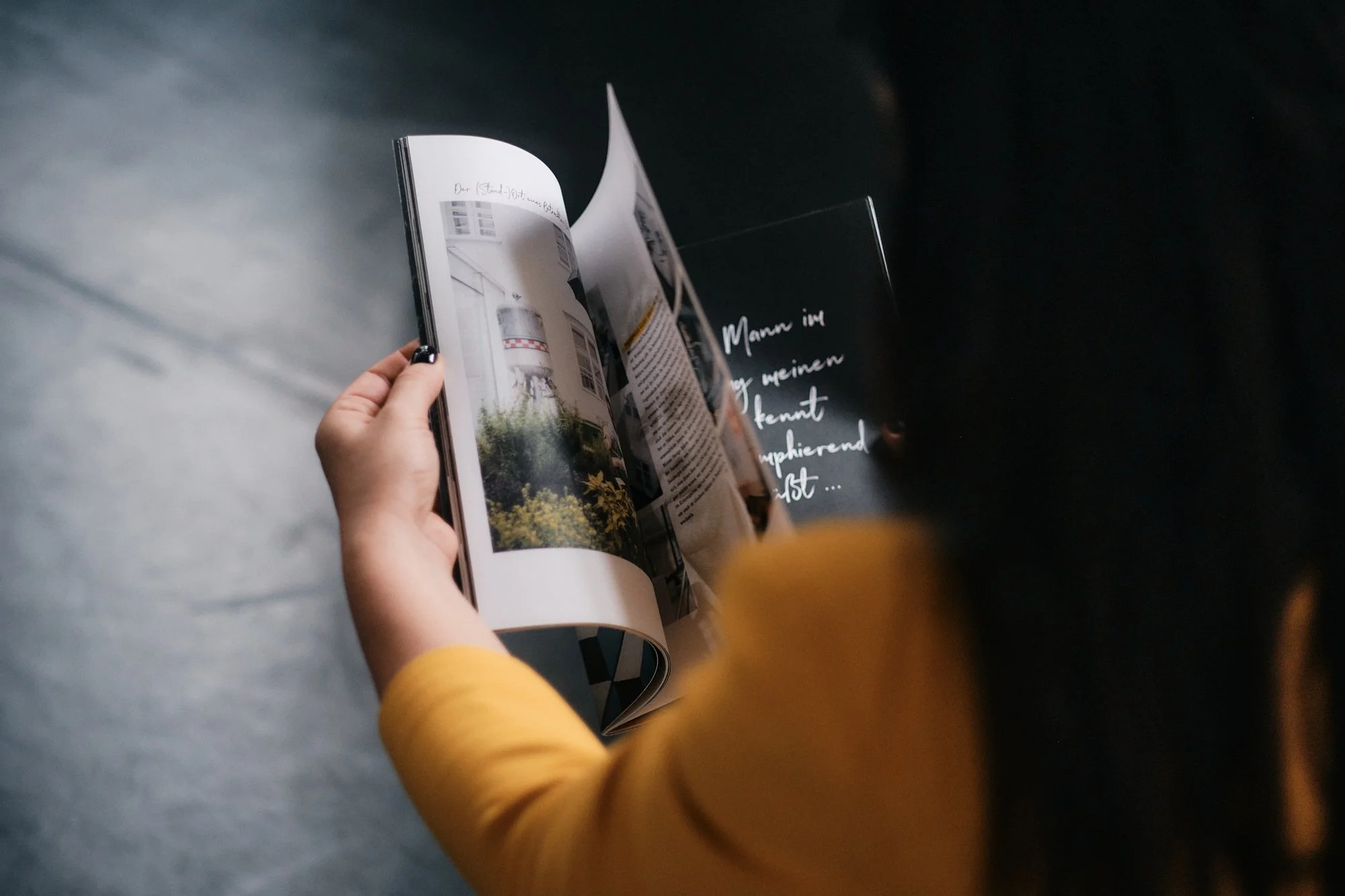 Eine Person mit gelben Pullover liest ein Magazin. Das Magazin zeigt ein Gebäude mit einem Wasserbecken und Pflanzen im Außenbereich. Es ist dunkel, und es sind weiße handschriftliche notizen auf schwarzen Hintergrund sichtbar.