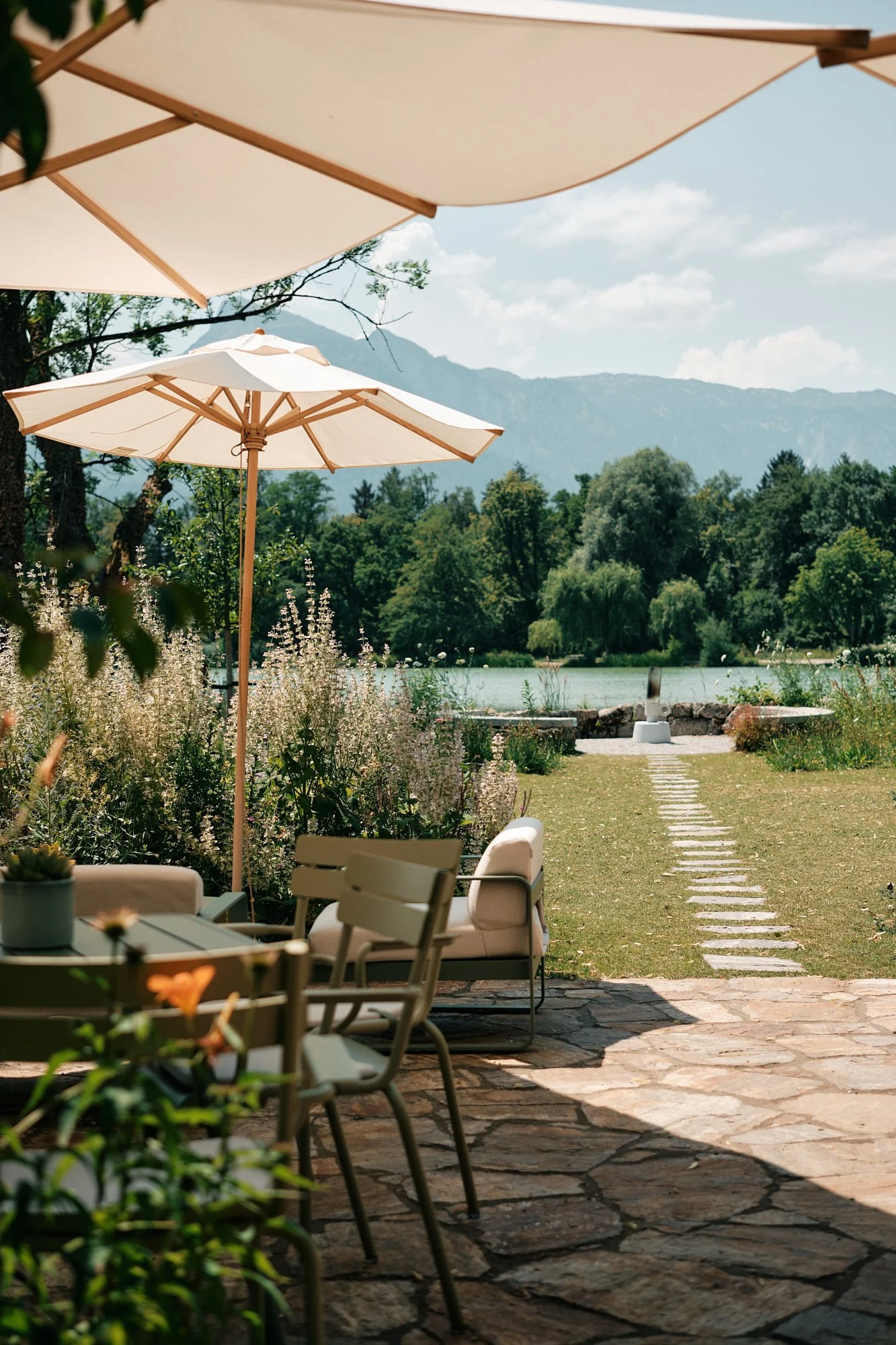 Ein ruhiger Garten mit Sitzmöbeln und Sonnenschirmen, Blick auf einen Fluss und Berge im Hintergrund.
