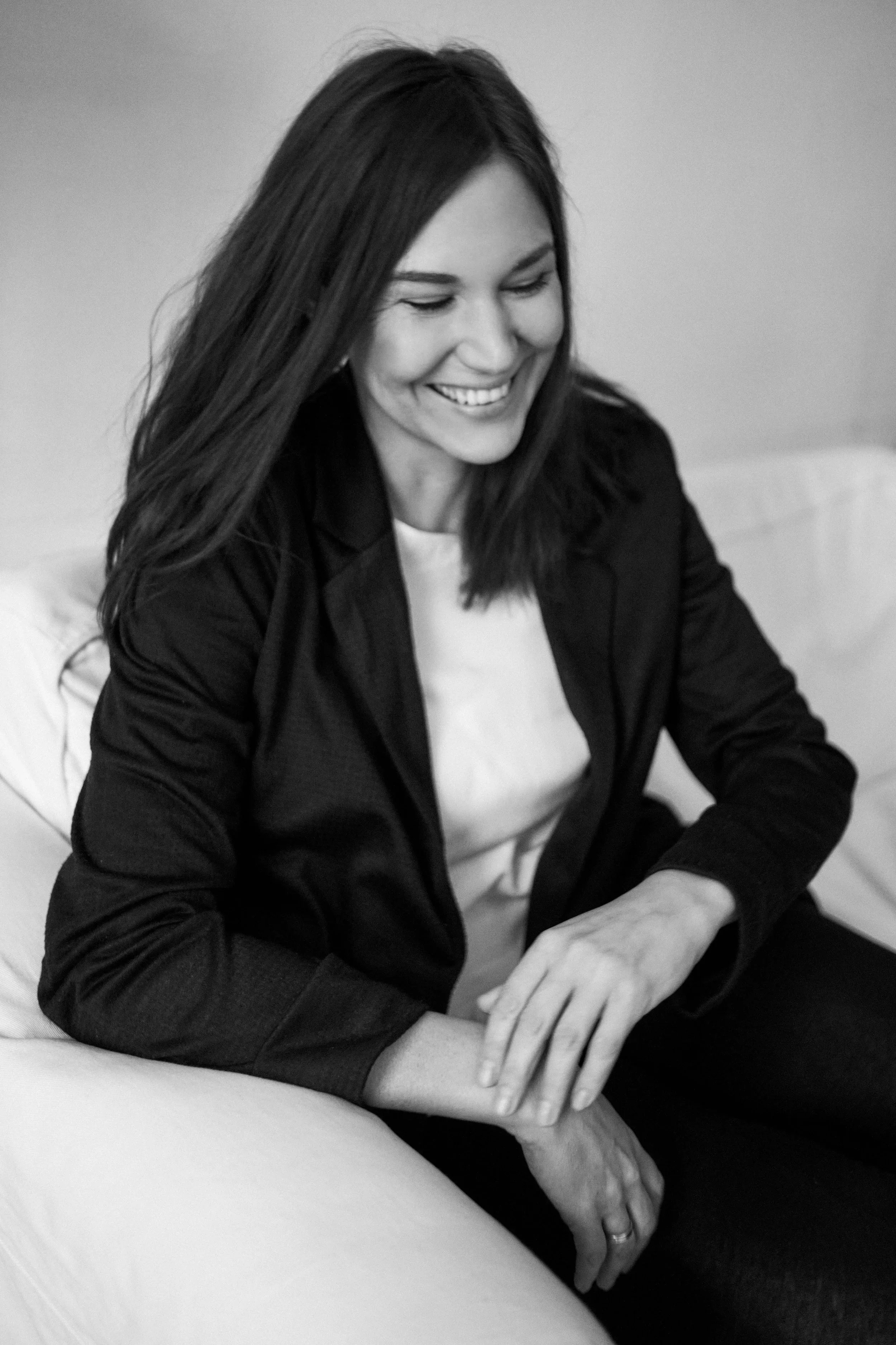 Schwarz-weiß-Foto einer lachenden Frau mit dunklem Haar, sitzend, mit einem weißen T-Shirt und einer dunklen Jacke, die in einer entspannten Haltung auf einem Sofa sitzt.