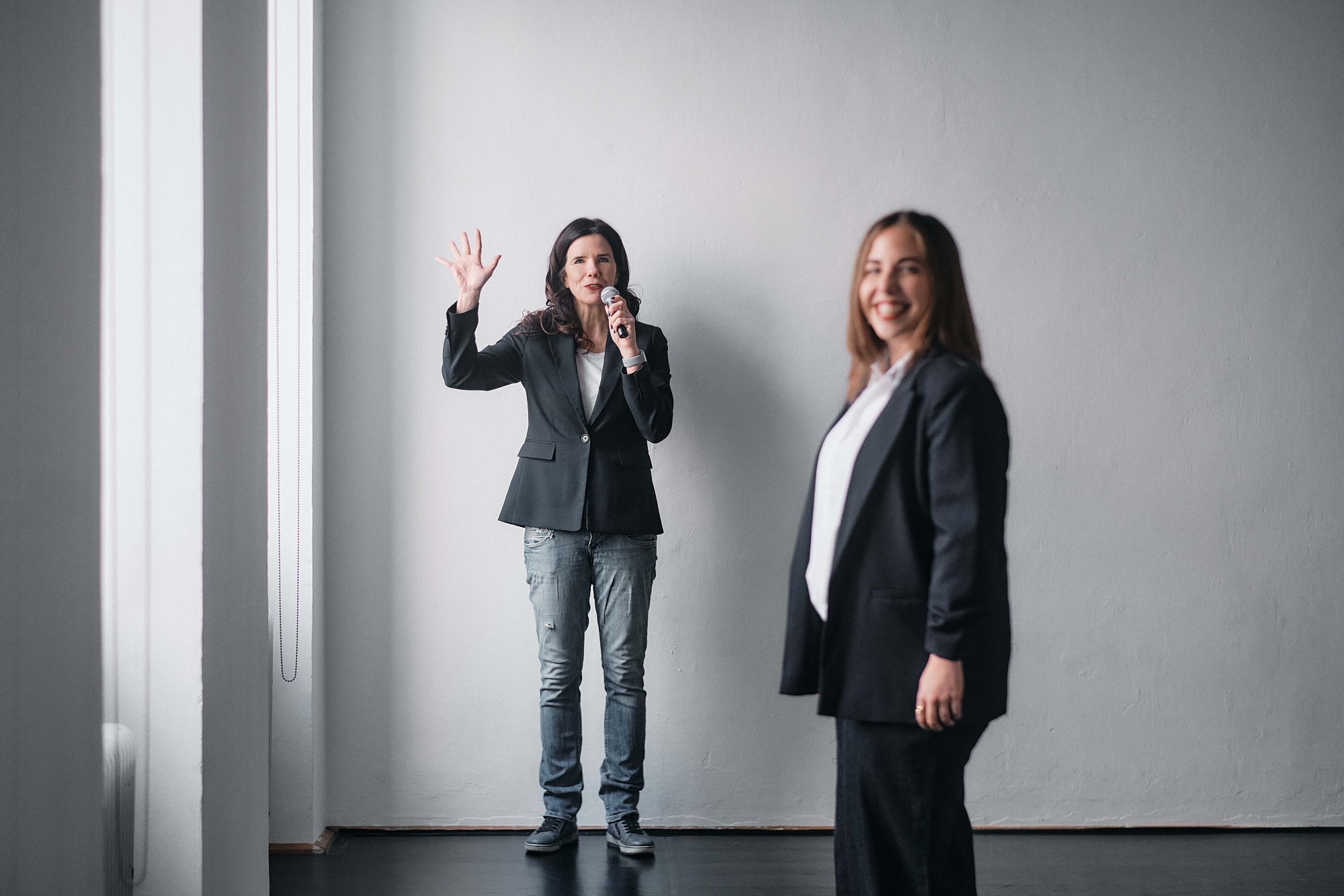 Zwei Frauen in businessähnlicher Kleidung stehen in einem Raum mit weißer Wand. Die Frau im Hintergrund hält ein Mikrofon und winkt, während die andere Frau im Vordergrund lächelt.