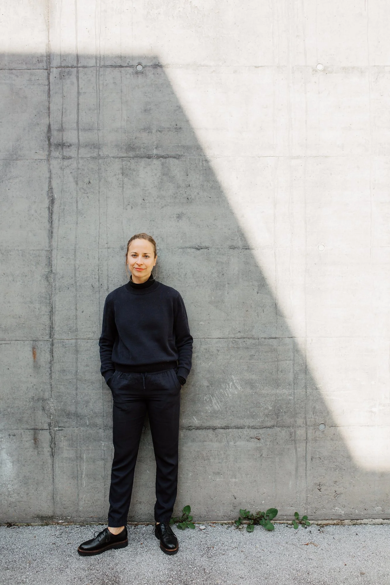 Junge Frau in schwarzer Kleidung steht vor einer grauen Betonwand im Schatten, mit Händen in den Taschen und einem leichten Lächeln.
