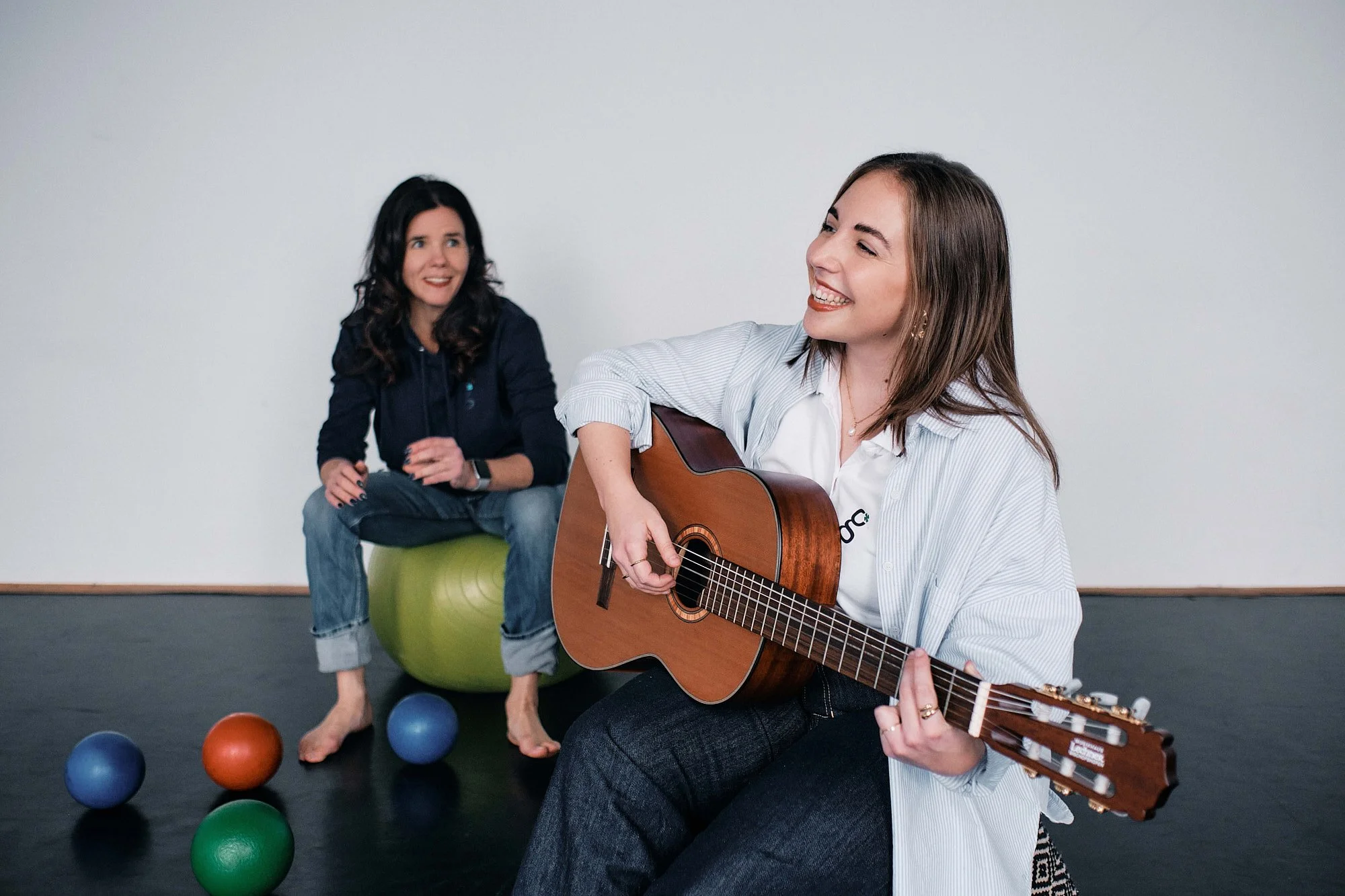 Zwei Frauen sitzen auf einem Gymball im Raum. Eine spielt Gitarre und beide lachen und haben Spaß.