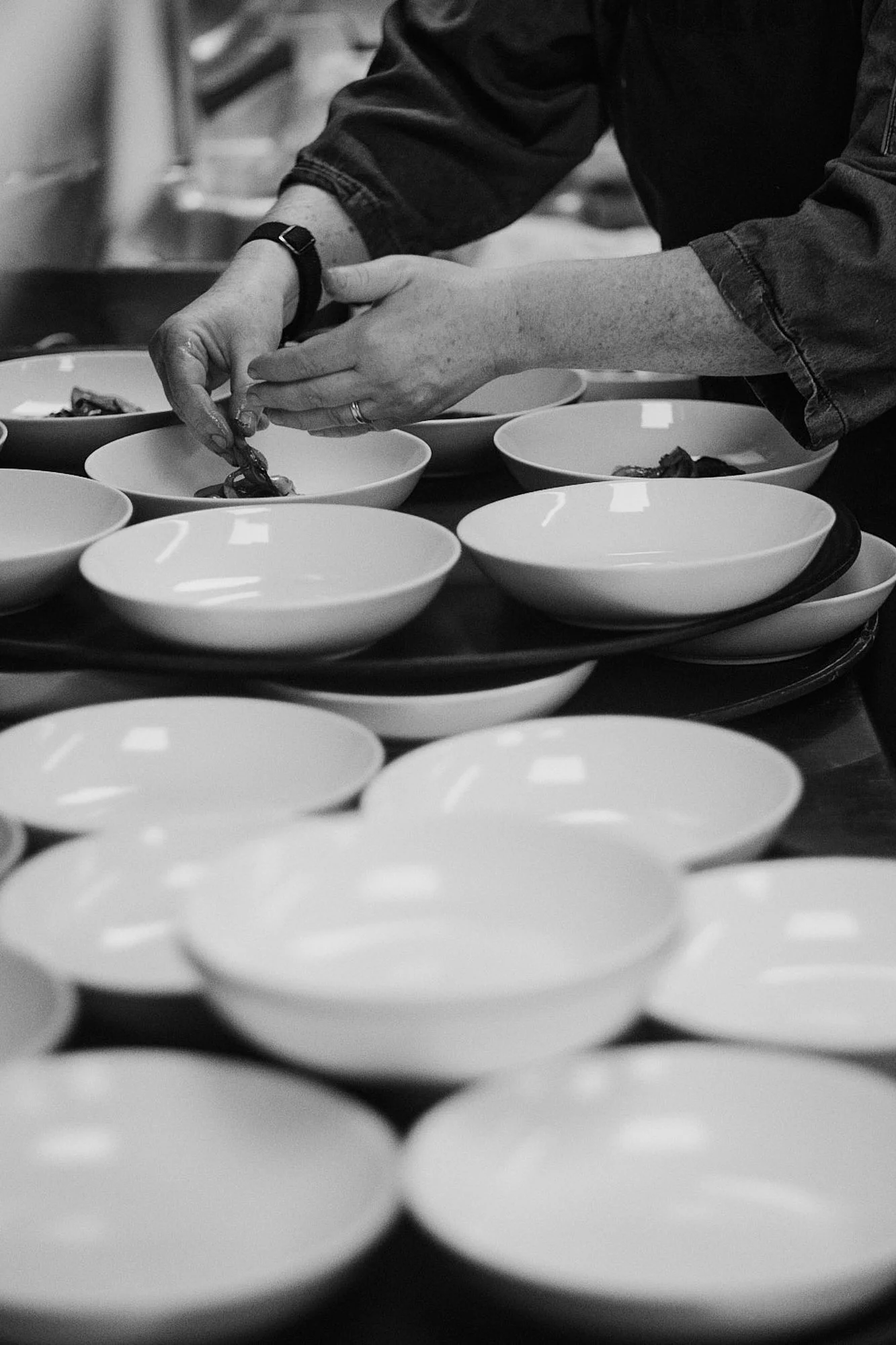 In einem schwarzen und weißen Bild sieht man eine Person, die in einer Küche am Kochen ist. Die Person ist dabei, Essen in kleine Schüsseln zu portionieren, die auf einem Tablett stehen. Die Person trägt eine Armbanduhr und ein langärmeliges Kleidung