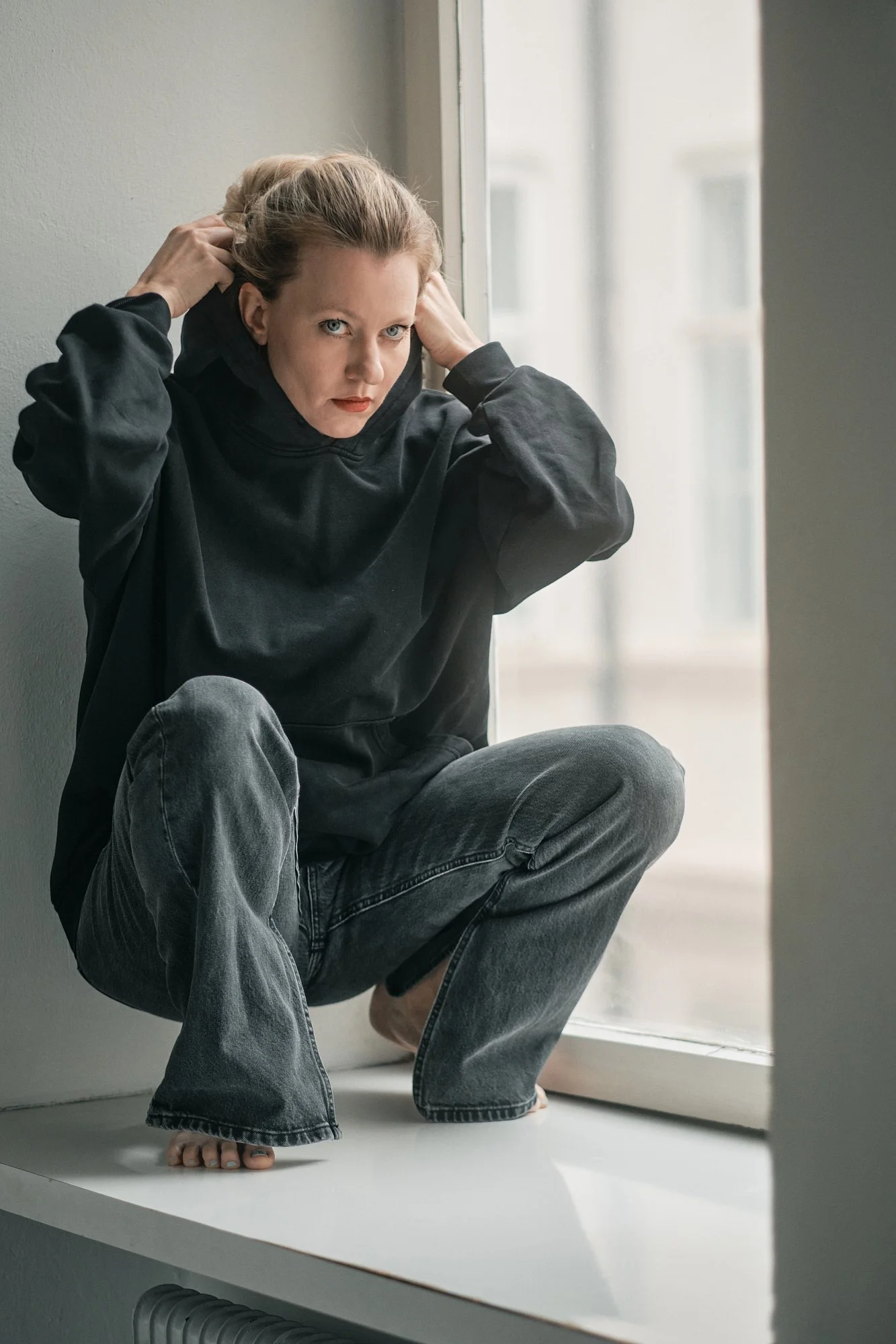 Ein Mädchen sitzt auf einer Fensterbank, trägt einen schwarzen Hoodie und alte Jeans, hat Dreadlocks und schaut direkt in die Kamera.