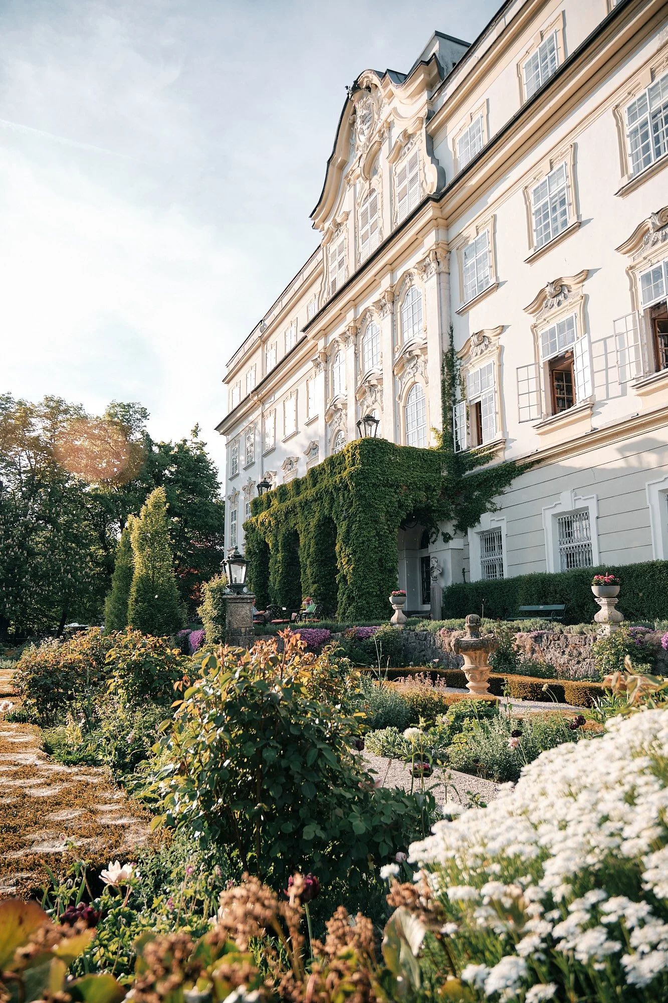 Historisches Gebäude mit barocker Fassade und vielen Fenstern, eingebettet in einen gepflegten Garten mit Brunnen, Bäumen und bunten Blumen bei Sonnenlicht.