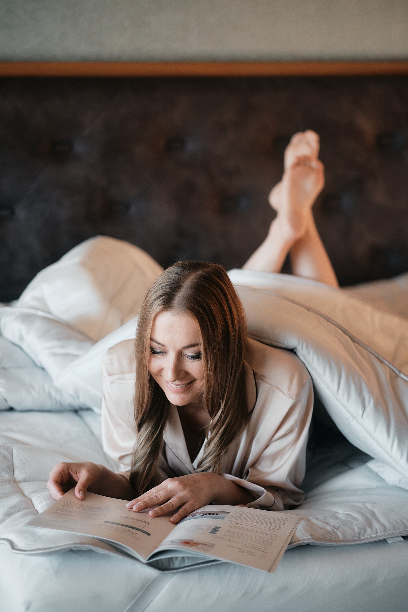 Eine lachende Frau mit braunen, langen Haaren liegt auf einem weißen Bett und liest eine Zeitschrift. Sie trägt eine beige Satin-Set mit einer schwarzen Wäsche darunter. Der Hintergrund ist eine dunkel gemusterte Wand, die nach oben hin sichtbar ist.