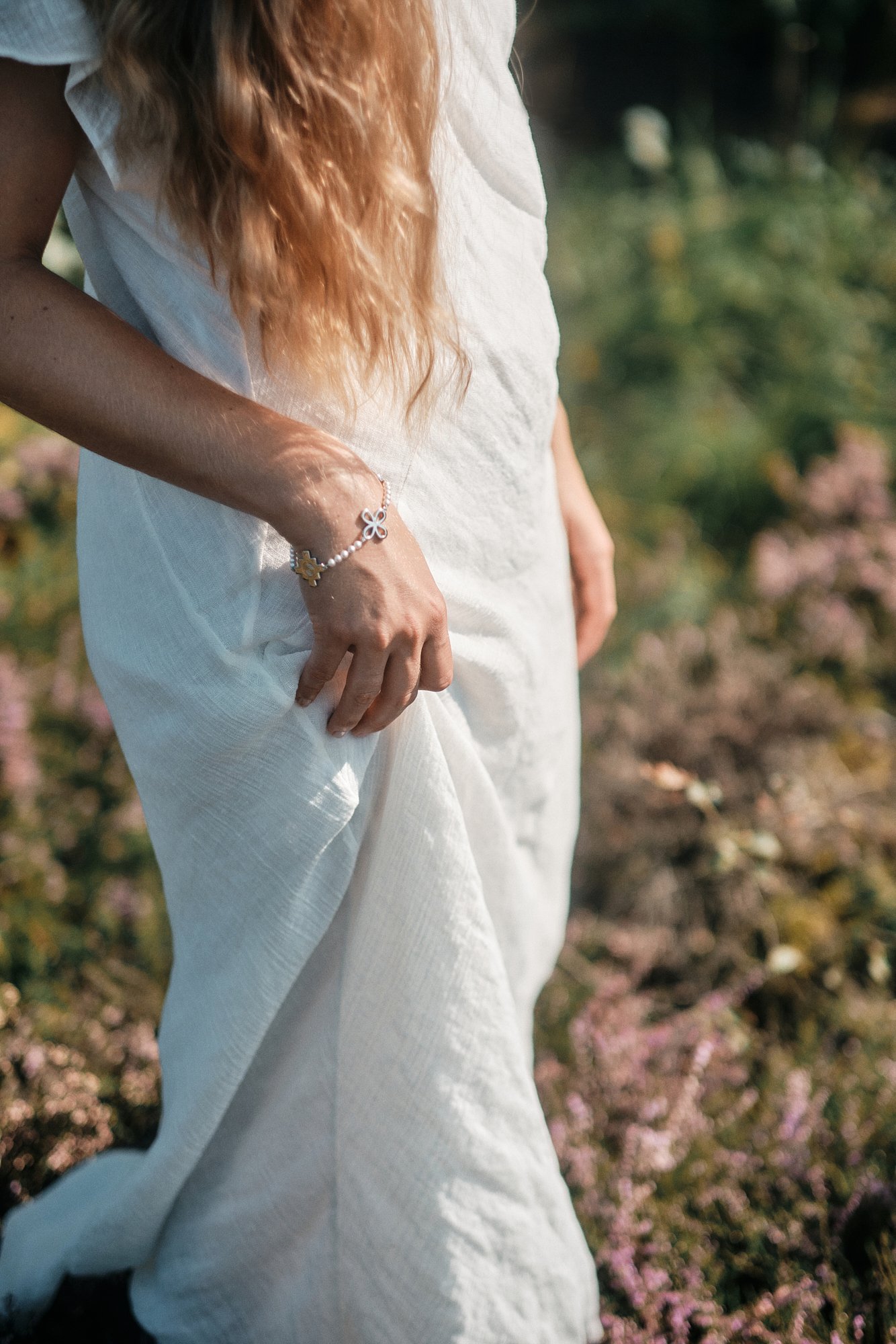 Nahaufnahme eines Menschen in weißem Kleid mit einer silbernen Armband in einer Blumenwiese im Sonnenlicht.