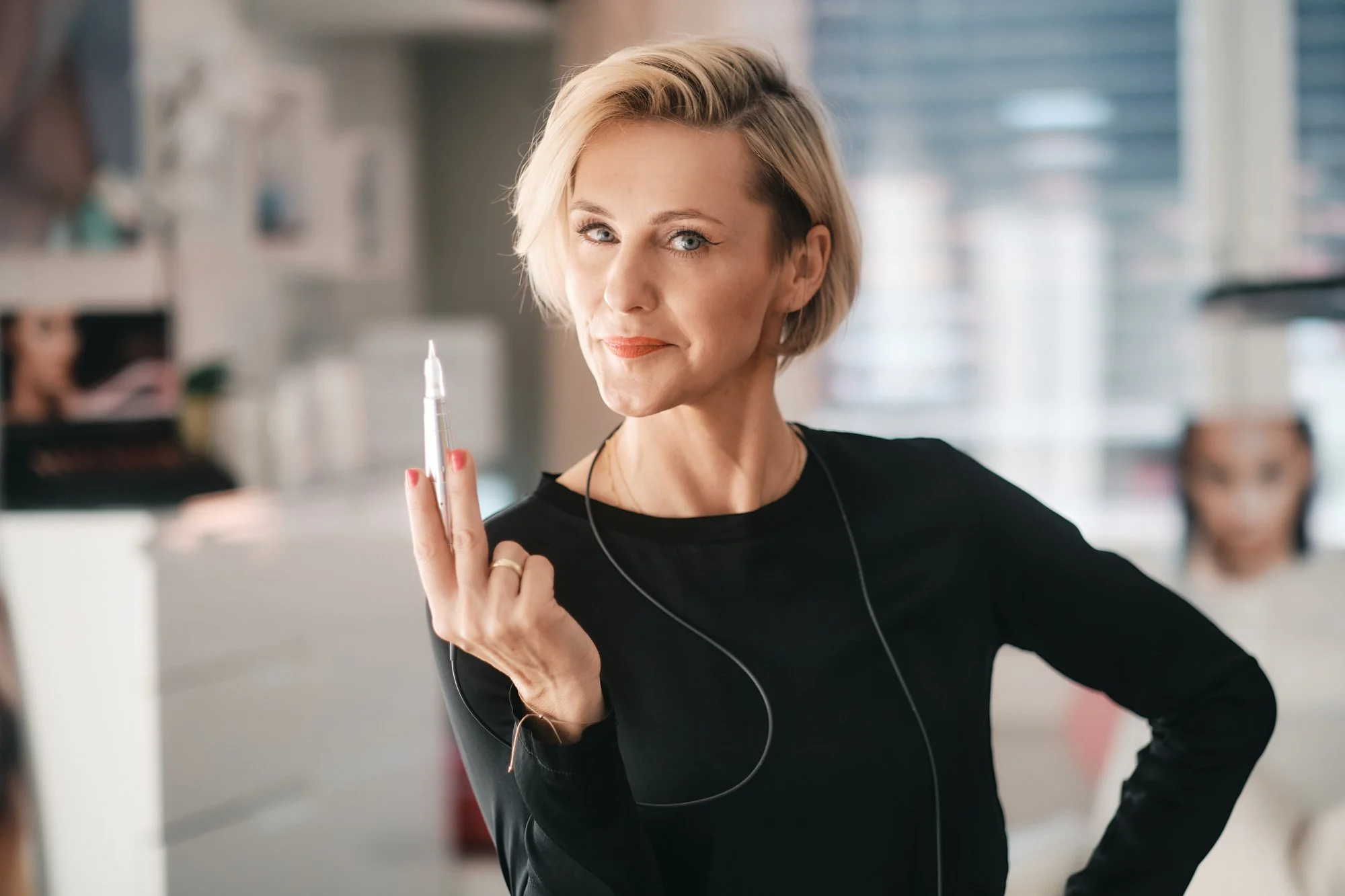 Frau mit Kurzhaarfrisur hält einen Kugelschreiber hoch, trägt eine schwarze Kleidung und hat eine selbstbewusste Ausstrahlung in einem modernen Büro.