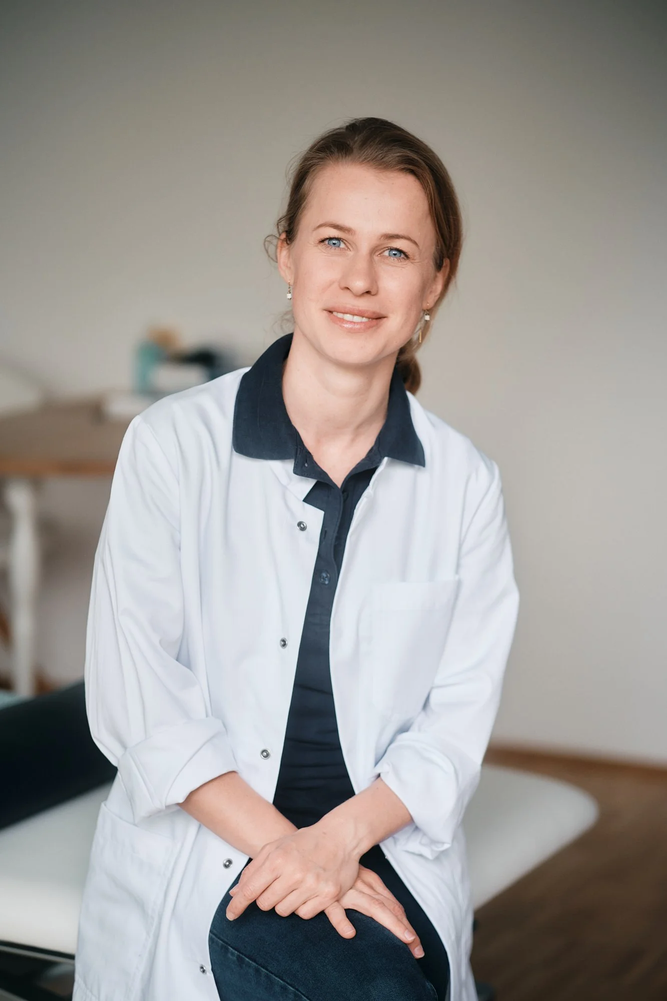 Eine freundliche Ärztin in Weißkrankenhausuniform sitzt auf einem Behandlungsstuhl in einer Praxis, schaut in die Kamera und lächelt. Im Hintergrund sind medizinische Geräte sichtbar.