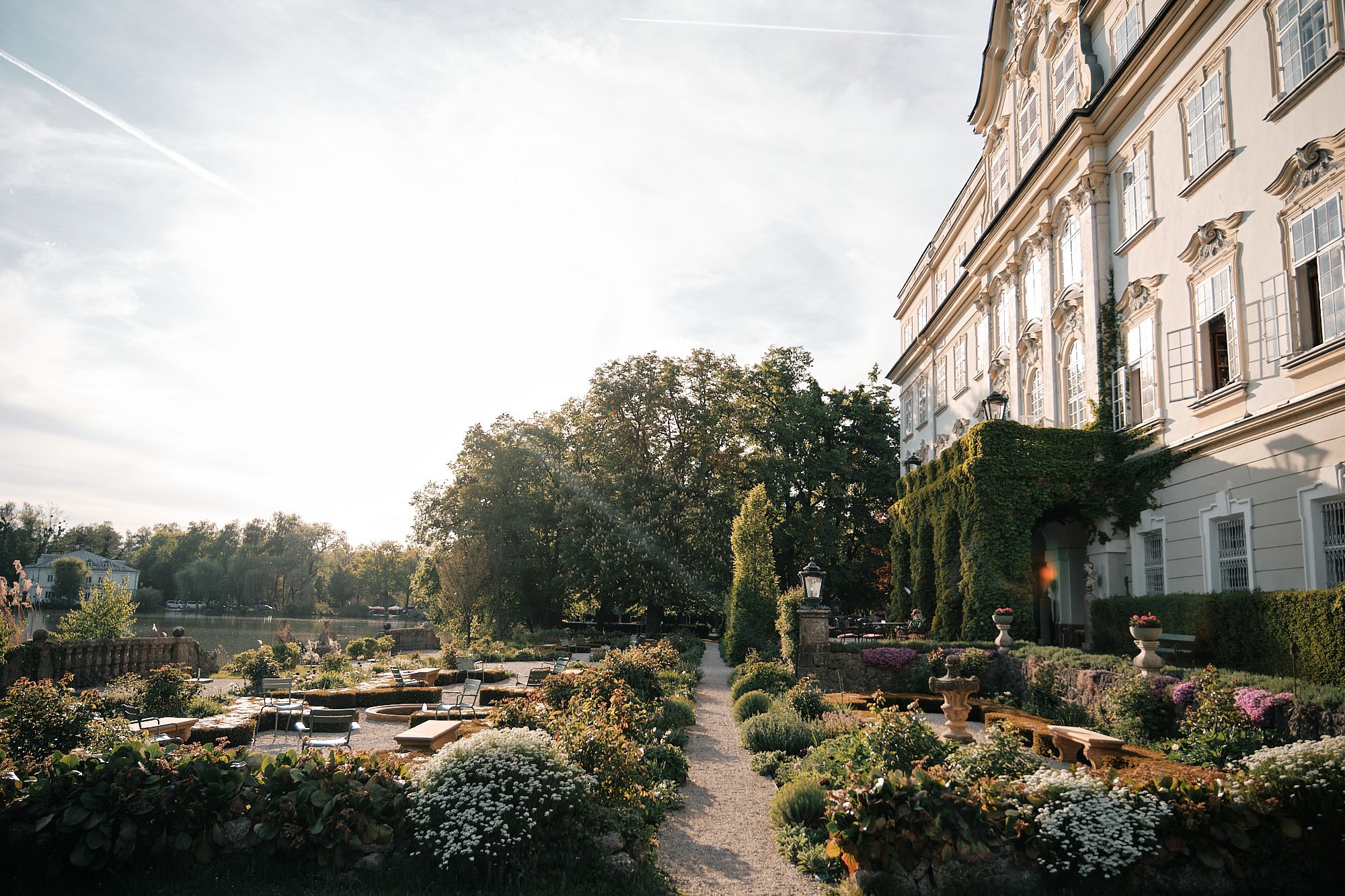 Ein großes, elegantes weißes Gebäude mit mehreren Fenstern, umgeben von einem gepflegten Garten mit bunten Blumen, Bäumen und einem Wasserweg, bei Sonnenuntergang.