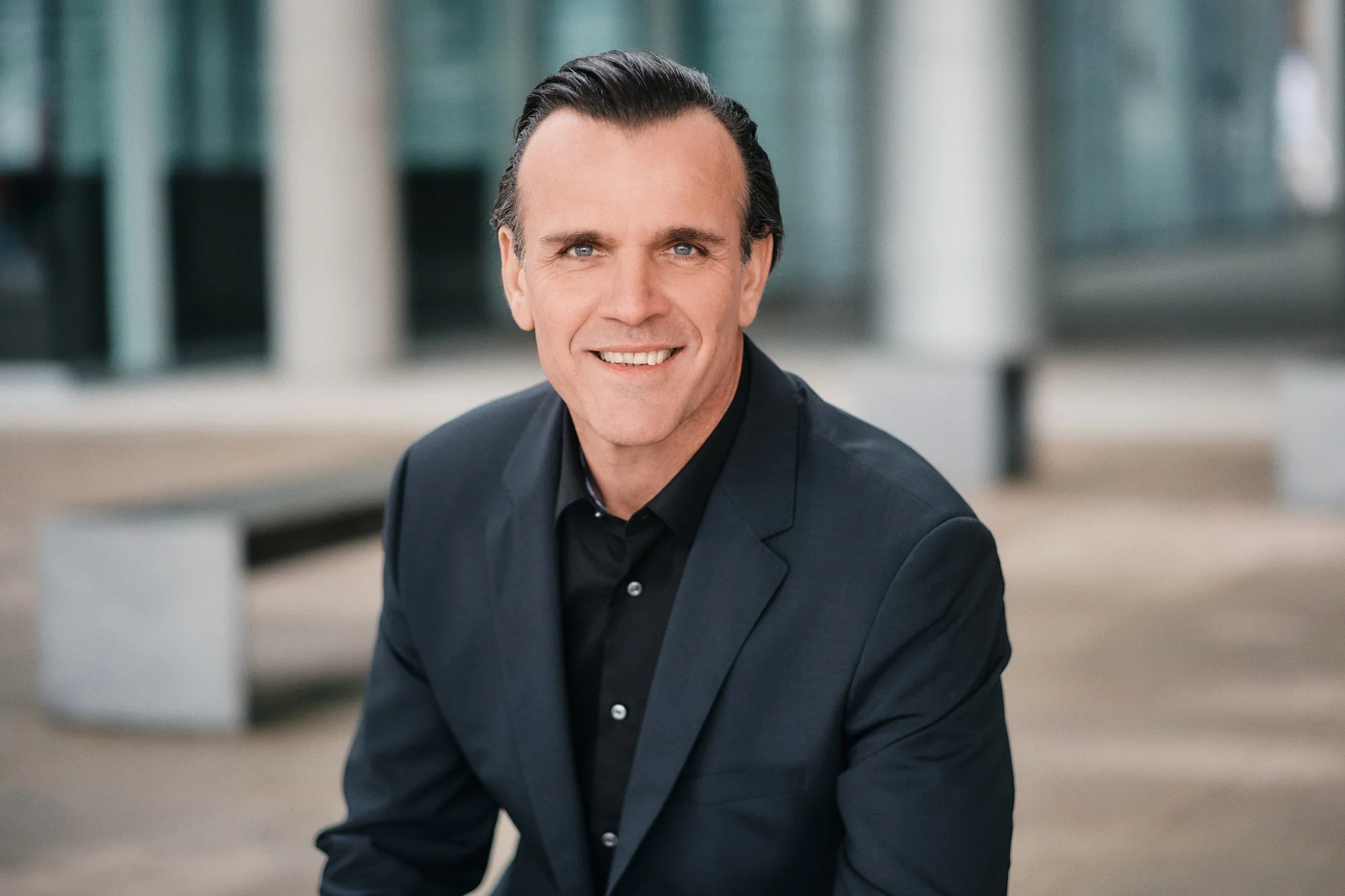 Ein Mann mit dunklem, kurz geschnittenem Haar trägt einen schwarzen Anzug und lächelt. Der Hintergrund ist unscharf und zeigt moderne Architektur im Freien.