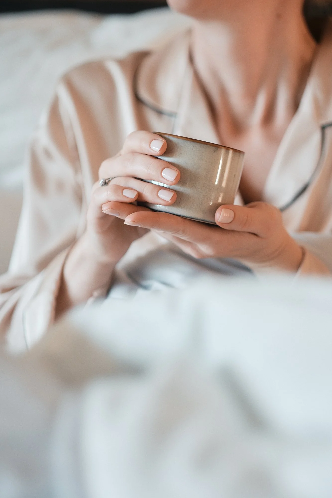 Nahaufnahme einer Person, die eine Tasse hält, im Hintergrund ein heller, seidiger Pyjama. Der Fokus liegt auf den Händen und der Tasse.