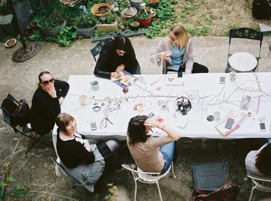 Fünf Frauen sitzen um eine rechteckige Esstafel im Garten. Es gibt Pflanzen im Hintergrund, und verschiedene Getränke sowie Handys auf dem Tisch.