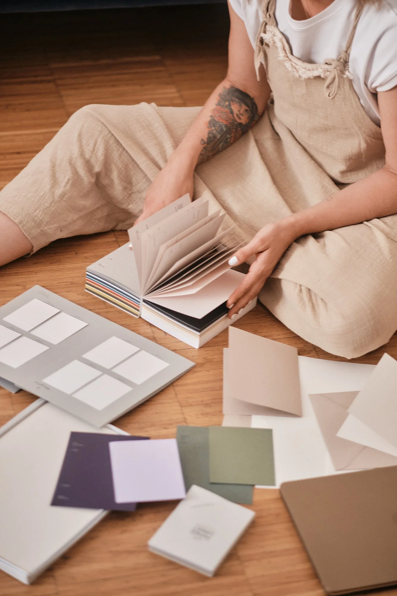 Eine Person sitzt auf dem Boden, liest in einem Farb- oder Materialmustersammelbuch und ist umgeben von Farb- und Materialproben auf dem Tisch. Die Person trägt helle Kleidung, hat eine Tätowierung am Arm und hält einen kleinen Farbkarte in der Hand.