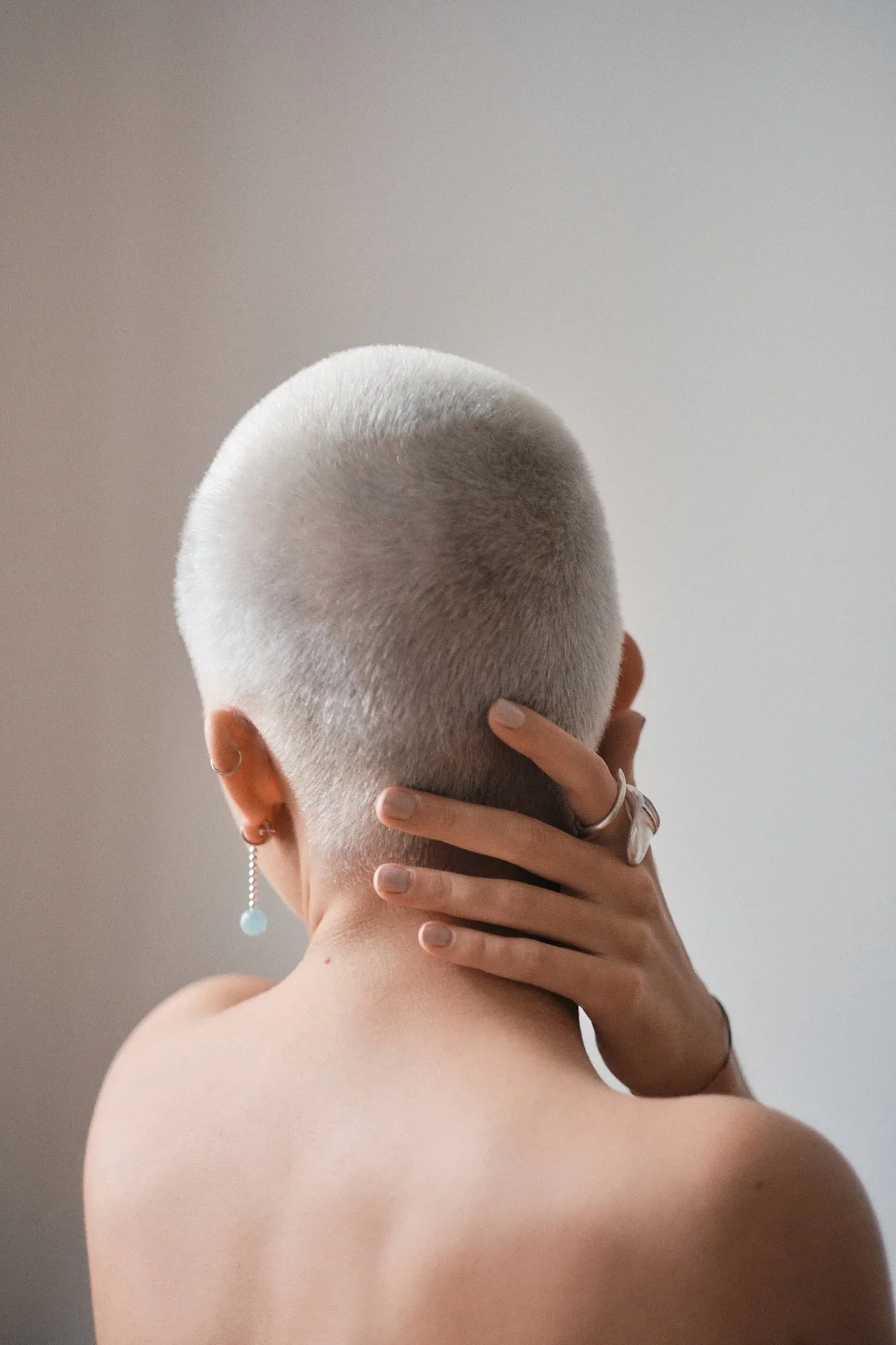 Nahaufnahme eines nackten Kopfes mit frisur, deren Seite rasiert ist, Person hält ihren Kopf mit einer Hand, trägt Schmuckohrringe und Ringe, im Hintergrund eine graue Wand.