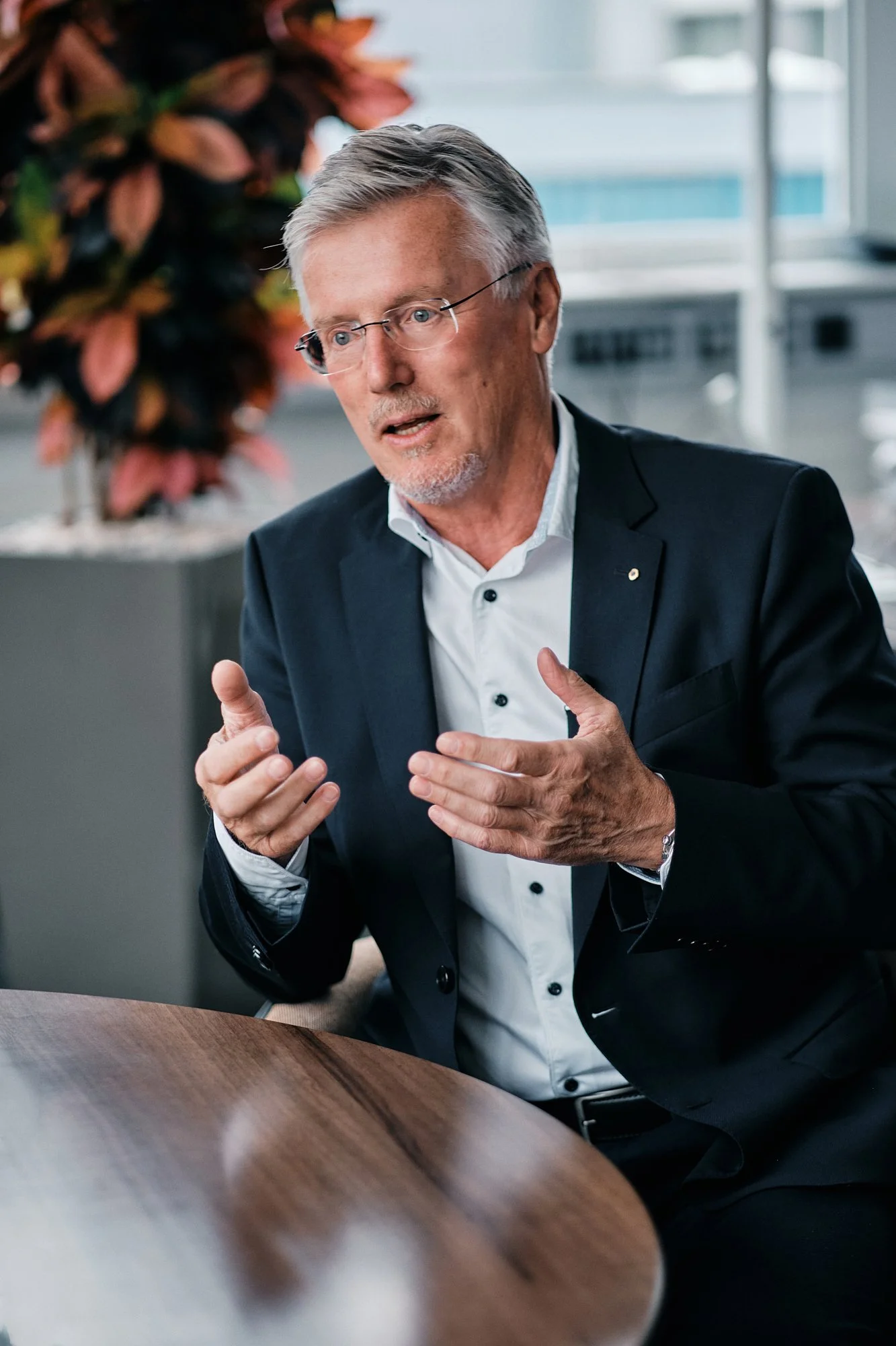 Ein älterer Mann in einem schwarzen Anzug und weißen Hemd sitzt an einem Tisch, spricht und gestikuliert während eines Gesprächs in einem modernen Büroumfeld, im Hintergrund große Fenster und Pflanzen.
