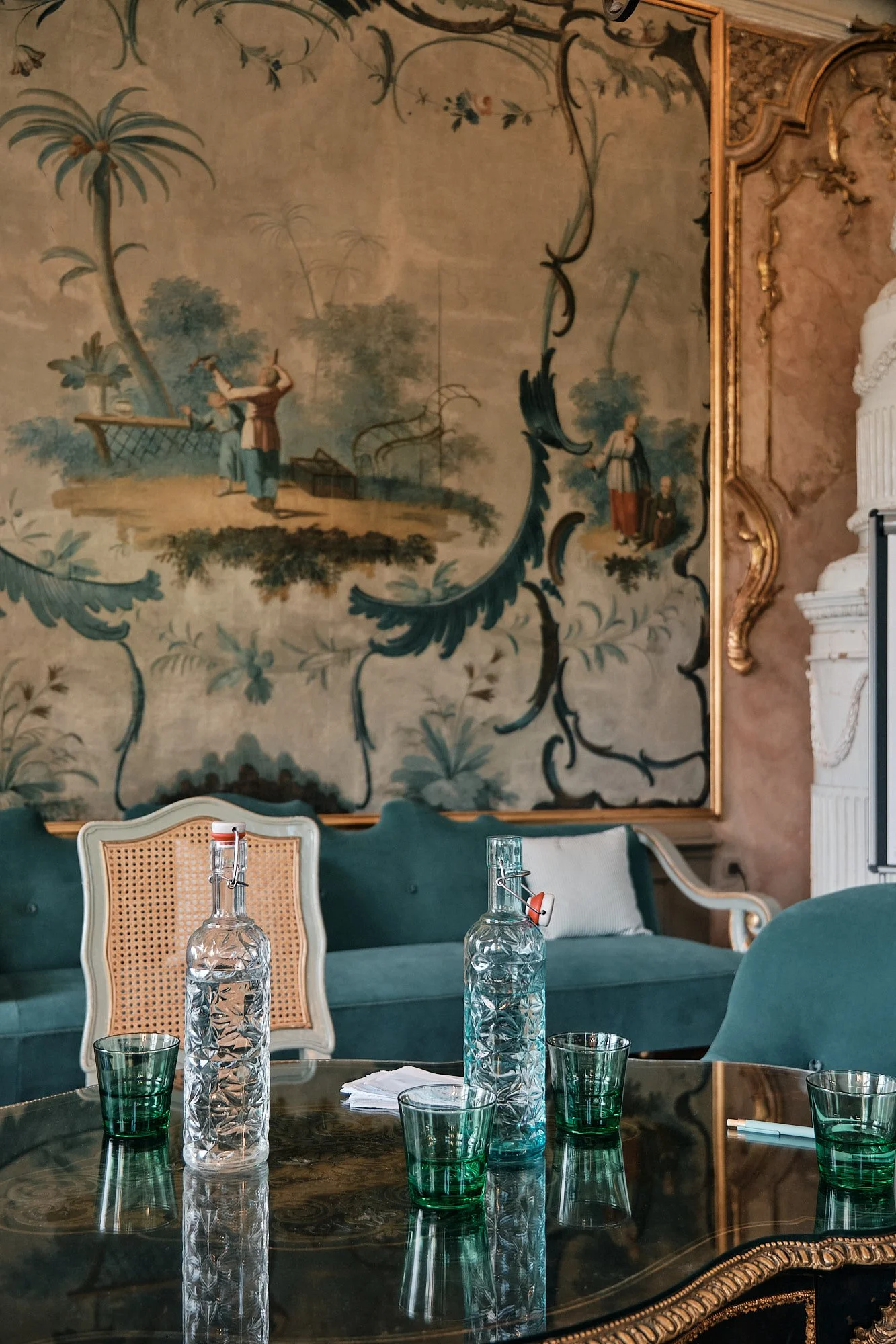 Ein eleganter Esszimmertisch mit sechs grünen Gläsern, zwei Wasserflaschen und Zettel. Im Hintergrund eine große Wandkunst mit einem Landschaftsmotiv und goldverzierten Rahmen sowie eine gemütliche Sitzbank