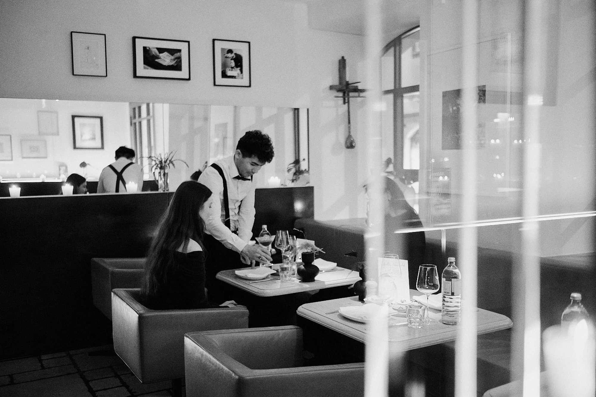 Ein schwarz-weiß Foto von einem Restaurant mit mehreren Personen. Zwei Kellner sind im Gespräch mit Gästen, während andere Gäste im Hintergrund an Tischen sitzen. Der Raum ist modern eingerichtet mit Kunstwerken an den Wänden, großen Fenstern und Ker
