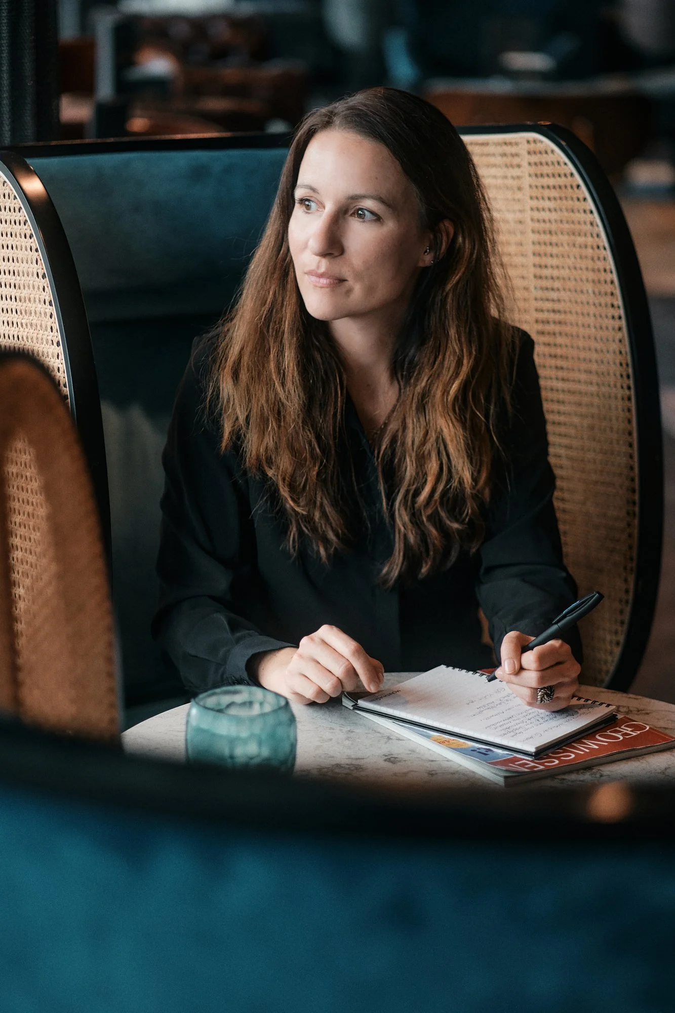Eine Frau mit langen braunen Haaren sitzt in einem Restaurant an einem runden Tisch, hält einen Stift in der Hand, hat ein Notizbuch und ein Magazin vor sich, und blickt nach links, während sie nachdenklich aussieht.