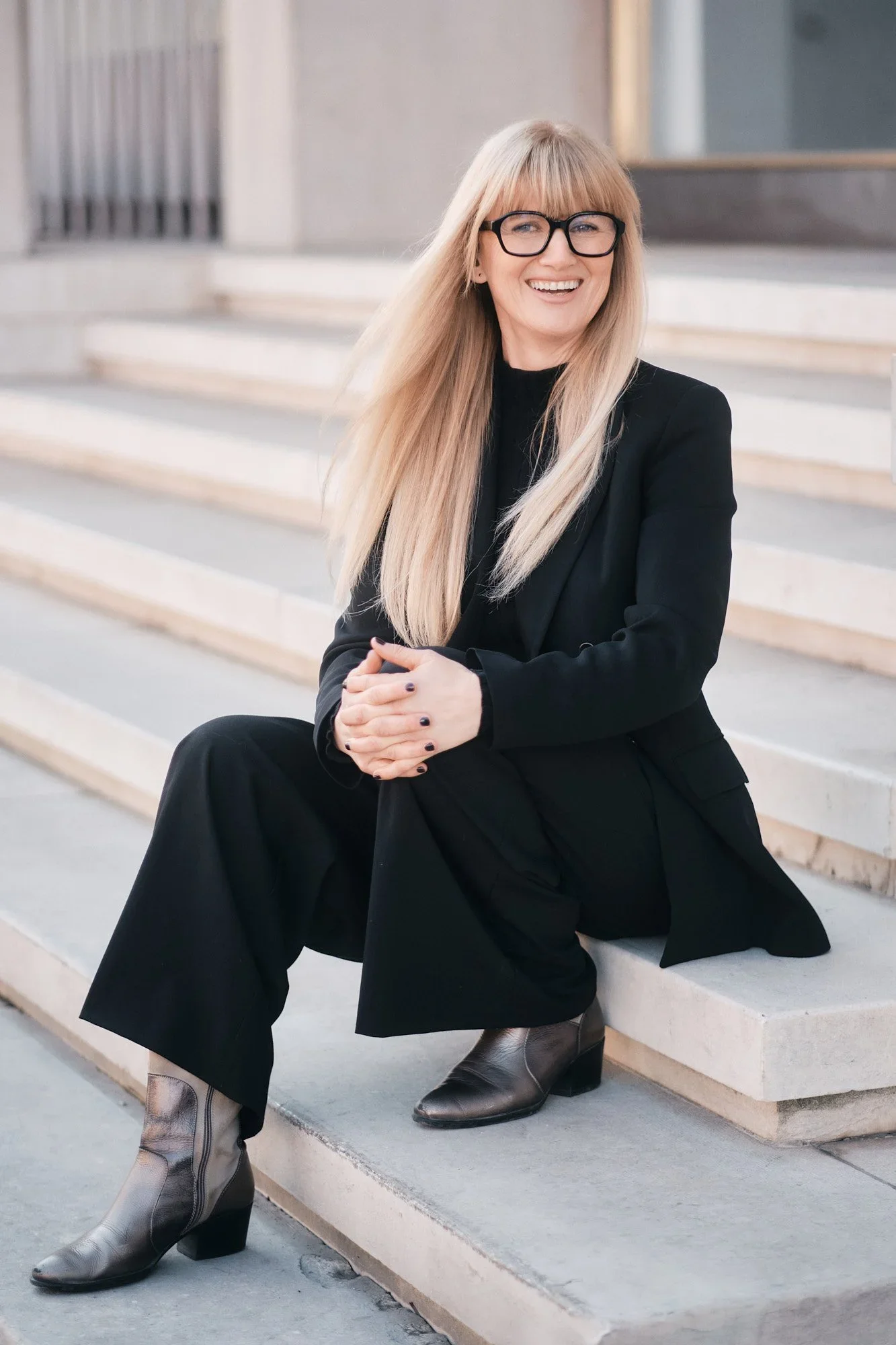 Eine lächelnde Frau mit langen blondem Haar, schwarzer Brille, schwarzem Blazer und Hose, sitzend auf einer Treppe aus hellem Stein, trägt graue Stiefel, im Hintergrund ein modernes Gebäude.
