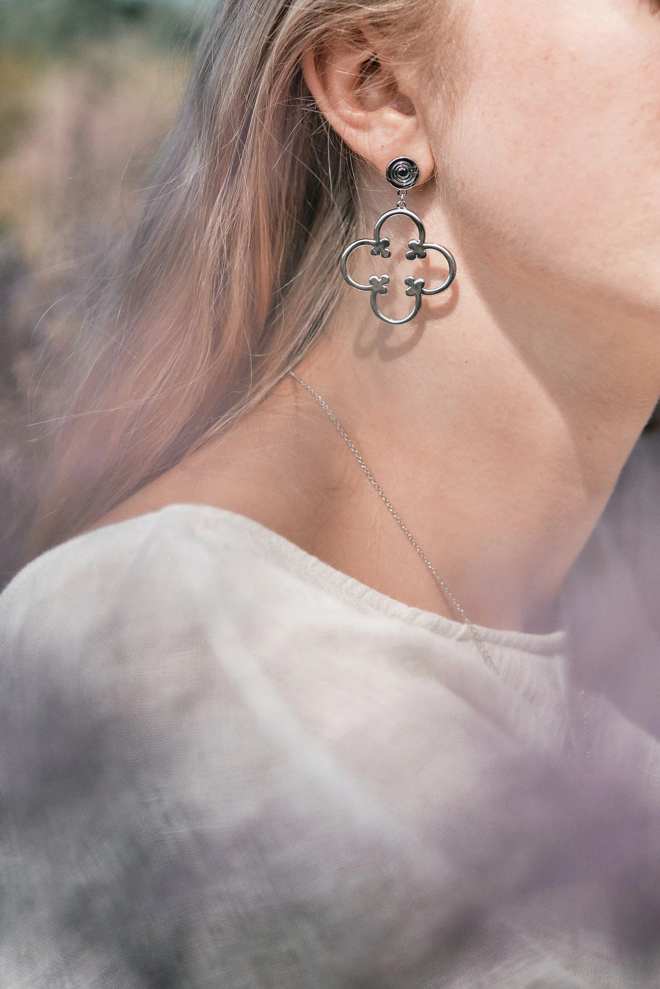 Nahaufnahme eines Ohrs mit einer großen silbernen Ohrring in Form eines vierblättrigen Kleeblattes und einem kleinen schwarzen Ohrpiercing, getragen von einer Person mit blonden Haaren und weißem Oberteil.