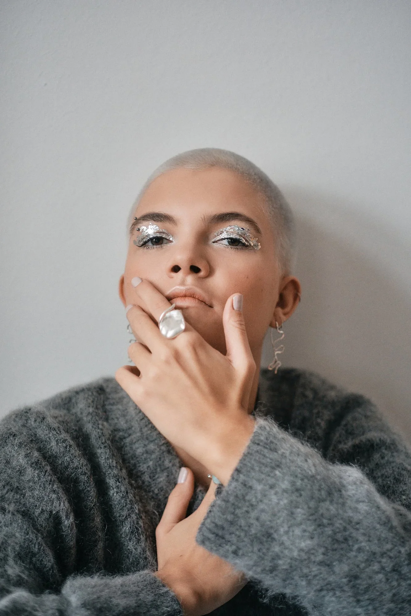 Porträt einer jungen Frau mit kurzgeschorenem Haar, auffälligem Make-up mit silberner Glitzer-Augenmakeup, die eine Hand mit mehreren Ringen vor dem Gesicht positioniert und einen grauen Wollpullover trägt.