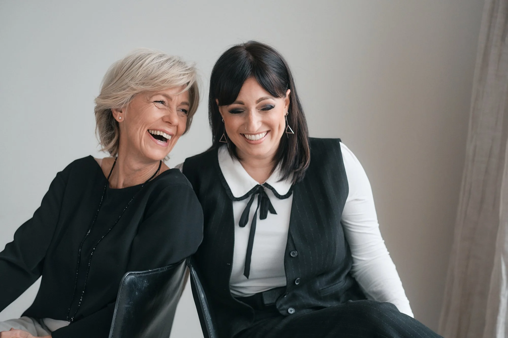 Zwei lachende Frauen, eine ältere und eine jüngere, sitzen gemeinsam und lachen. Sie tragen moderne Kleidung, die ältere Frau hat kurze blonde Haare, die jüngere schwarze Haare. Der Hintergrund ist neutral, wahrscheinlich eine weiße Wand.