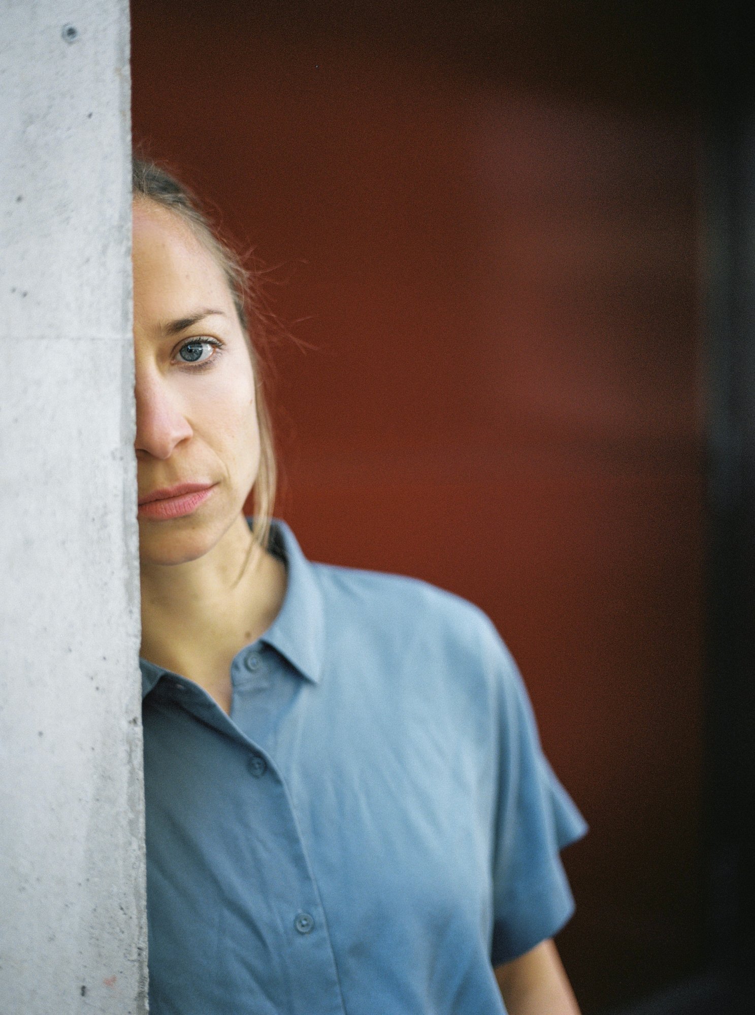 Nahaufnahme eines jungen Mädchens, das an einer weißen Wand lehnt. Sie trägt eine blaue Bluse und hat blonde Haare, schaut nach vorne, mit einem leichten Lächeln.