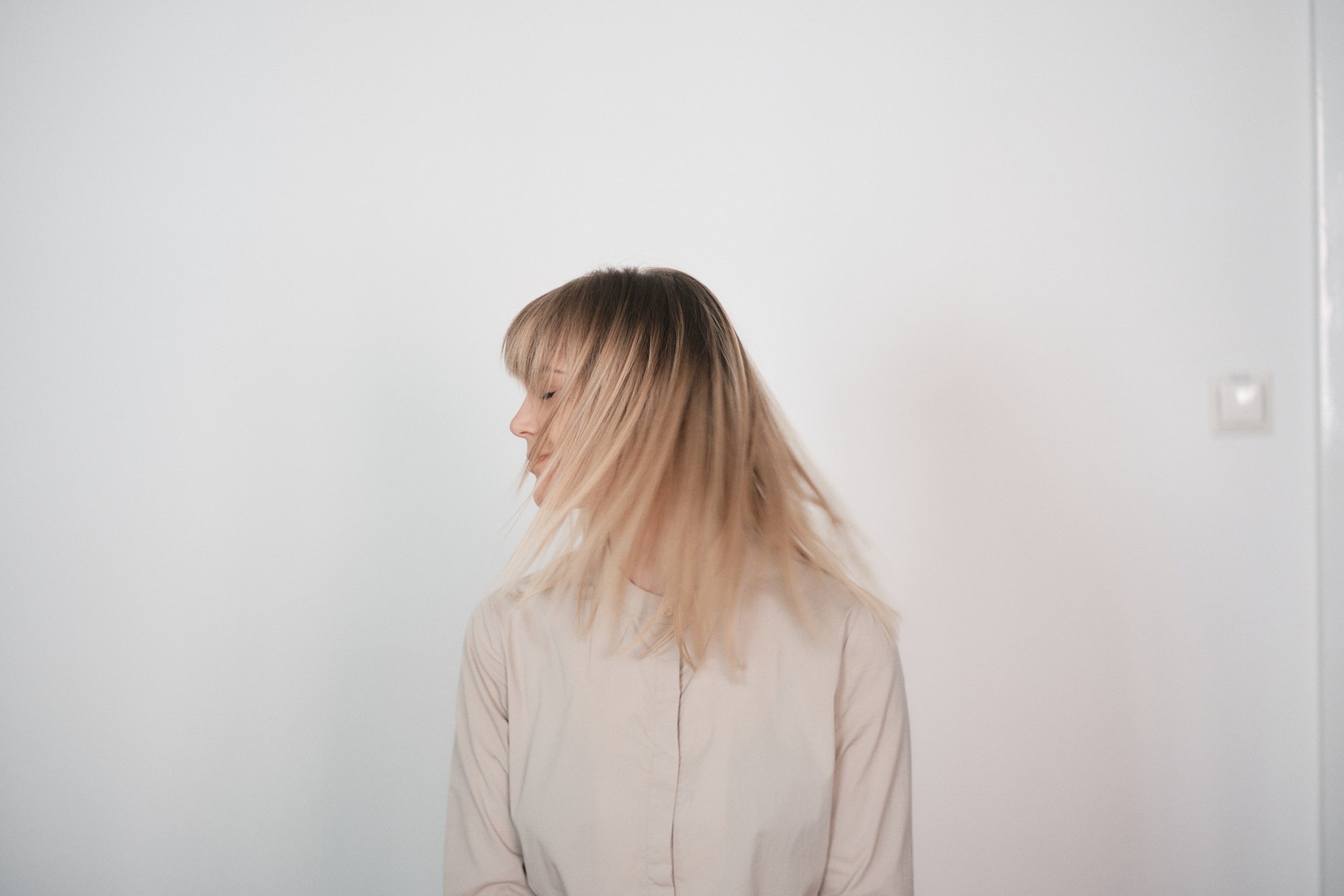 Ein junges Mädchen mit langen, blonden Haaren schaut nach links. Sie trägt eine helle Bluse und steht vor einer weißen Wand, auf der eine Steckdose zu sehen ist.