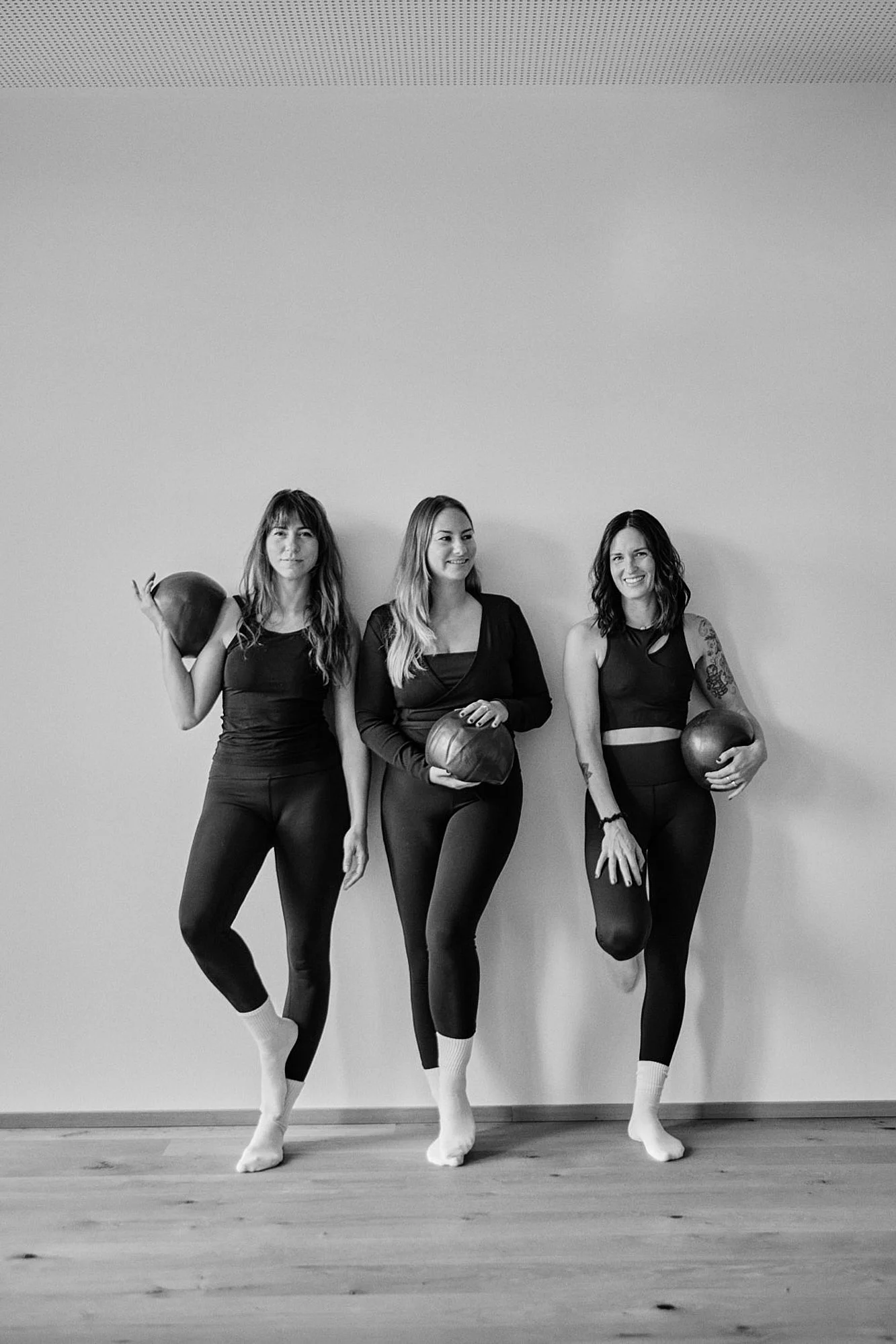 Drei Frauen in Sportkleidung und weißen Socken, die gegen eine helle Wand lehnen und Getränke in der Hand halten, in einem Raum mit Holzboden.