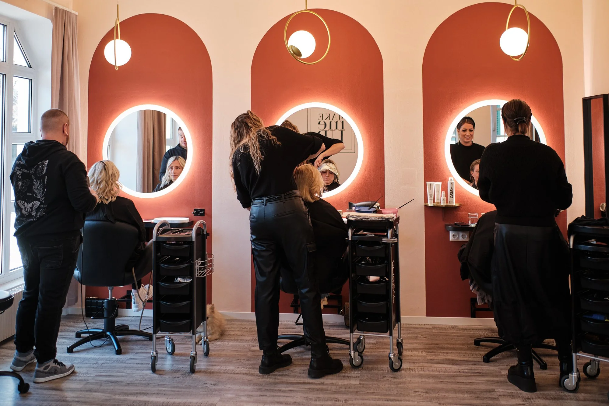 Menschen in einem Friseurstudio, einige sitzen beim Friseur, der einen Friseurhaarschnitt macht, während andere im Hintergrund mit Spiegeln und Arbeiten beschäftigt sind.