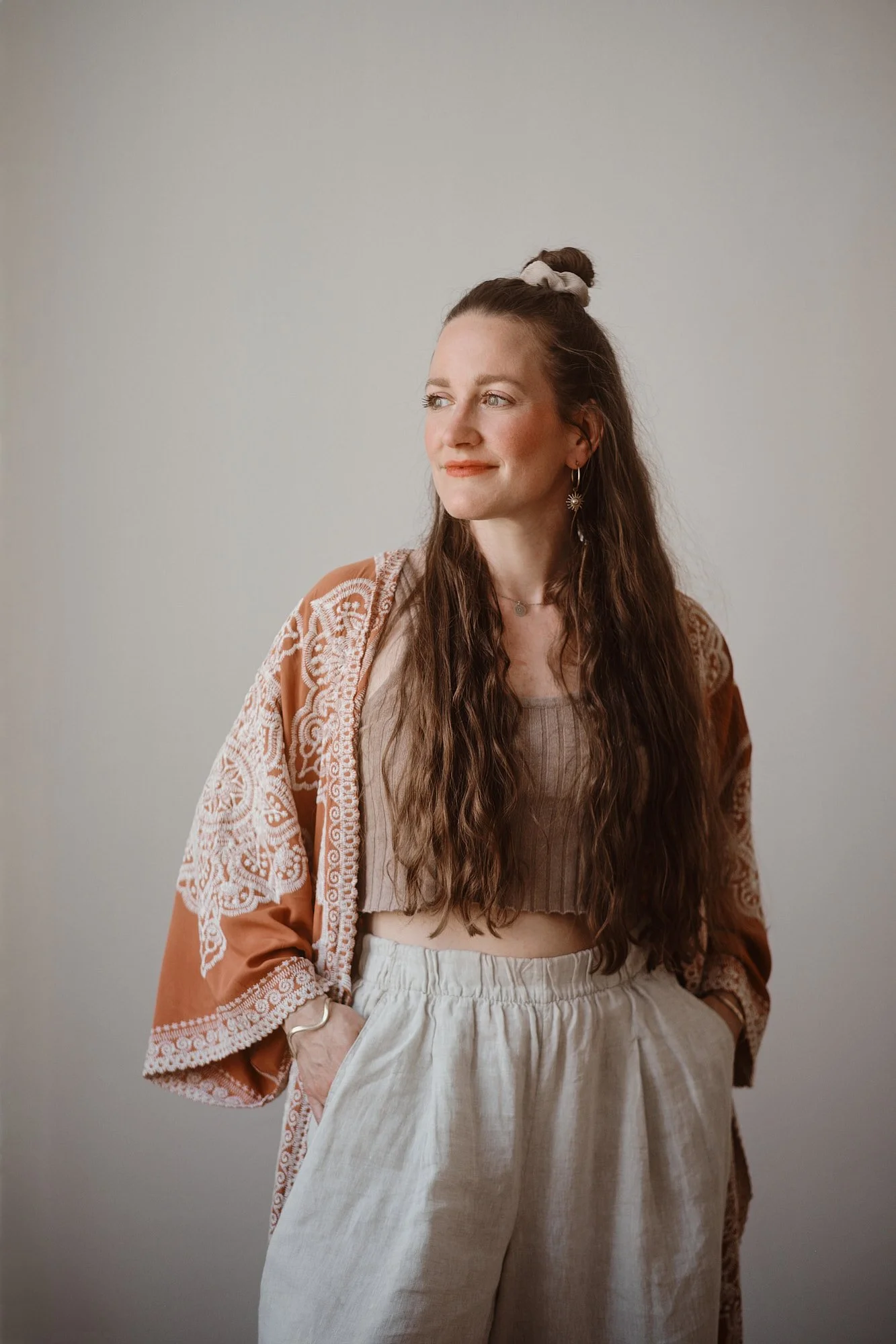 Frau mit langen brunswarmen Haaren, violetten Augen, trägt Ohrringe und einen braunen Kimono-Look, lehnt an der Wand, lächelt leicht, blickt zur Seite.