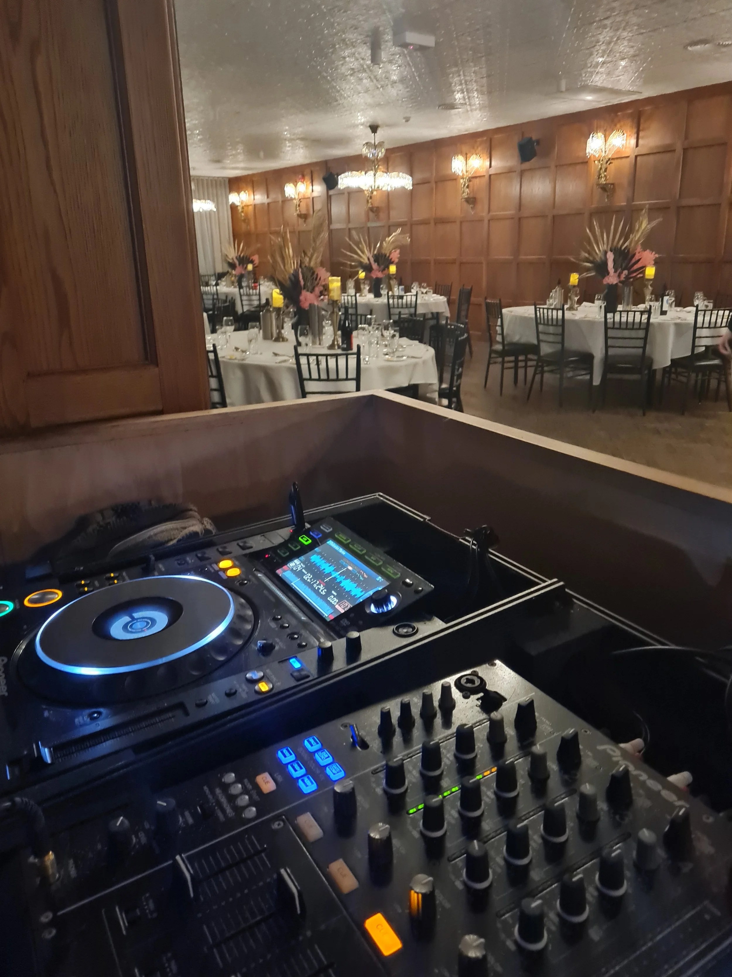 A DJ setup with turntables, a mixer, and a soundboard in a decorated banquet hall with tables and chairs, floral centerpieces, chandeliers, and wall sconces.