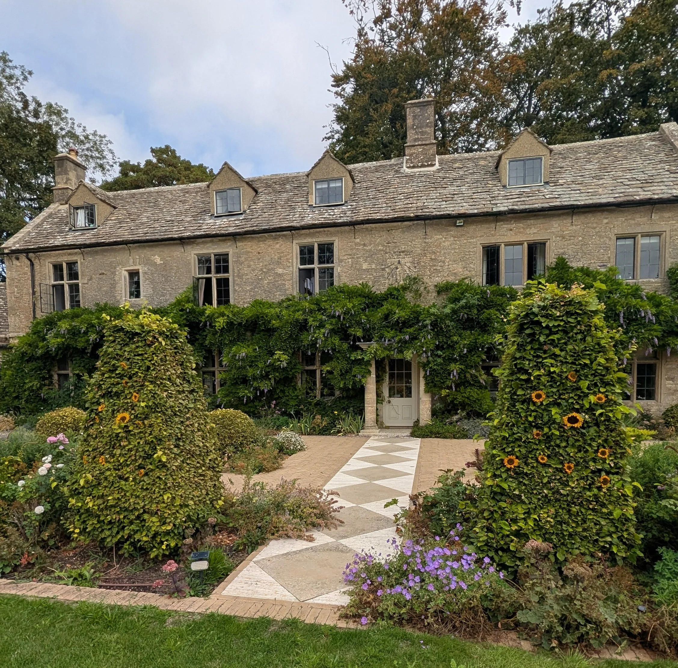 A charming stone house with a sloped roof, multiple chimneys, and small dormer windows. The house is surrounded by lush greenery, including colourful flowers, bushes, and trees, with a paved pathway leading up to the entry.