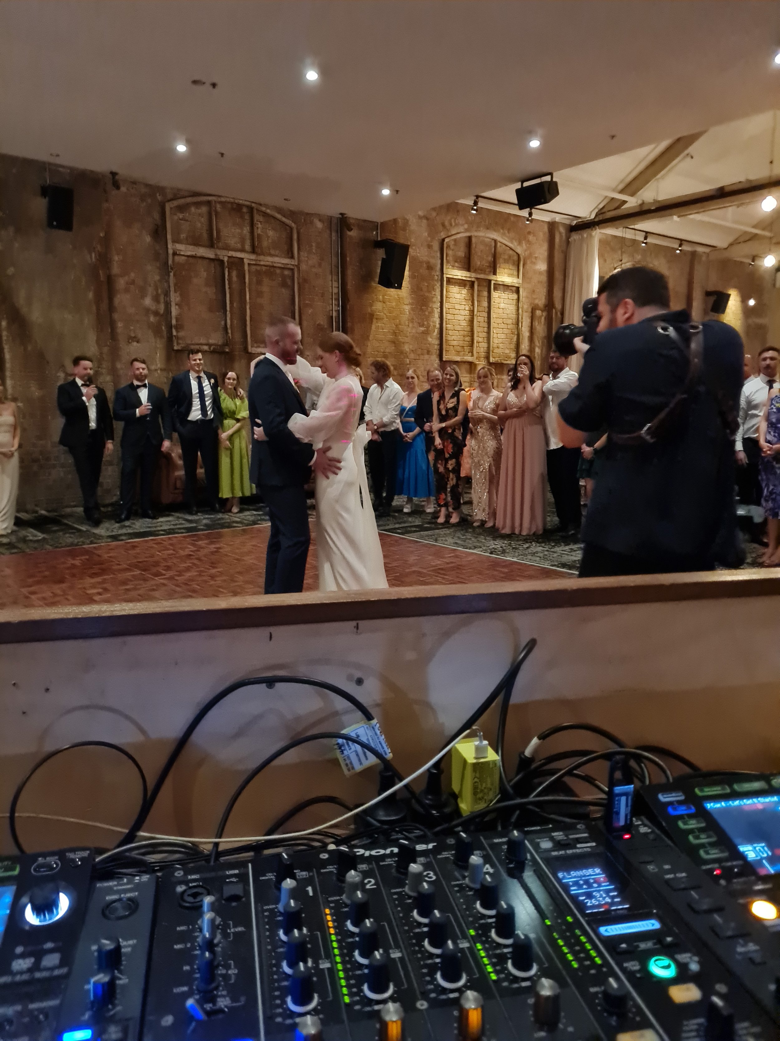 A DJ mixing console with various knobs, sliders, and colorful lights, and a DJ booth with a crowd in the background at a wedding reception or party.