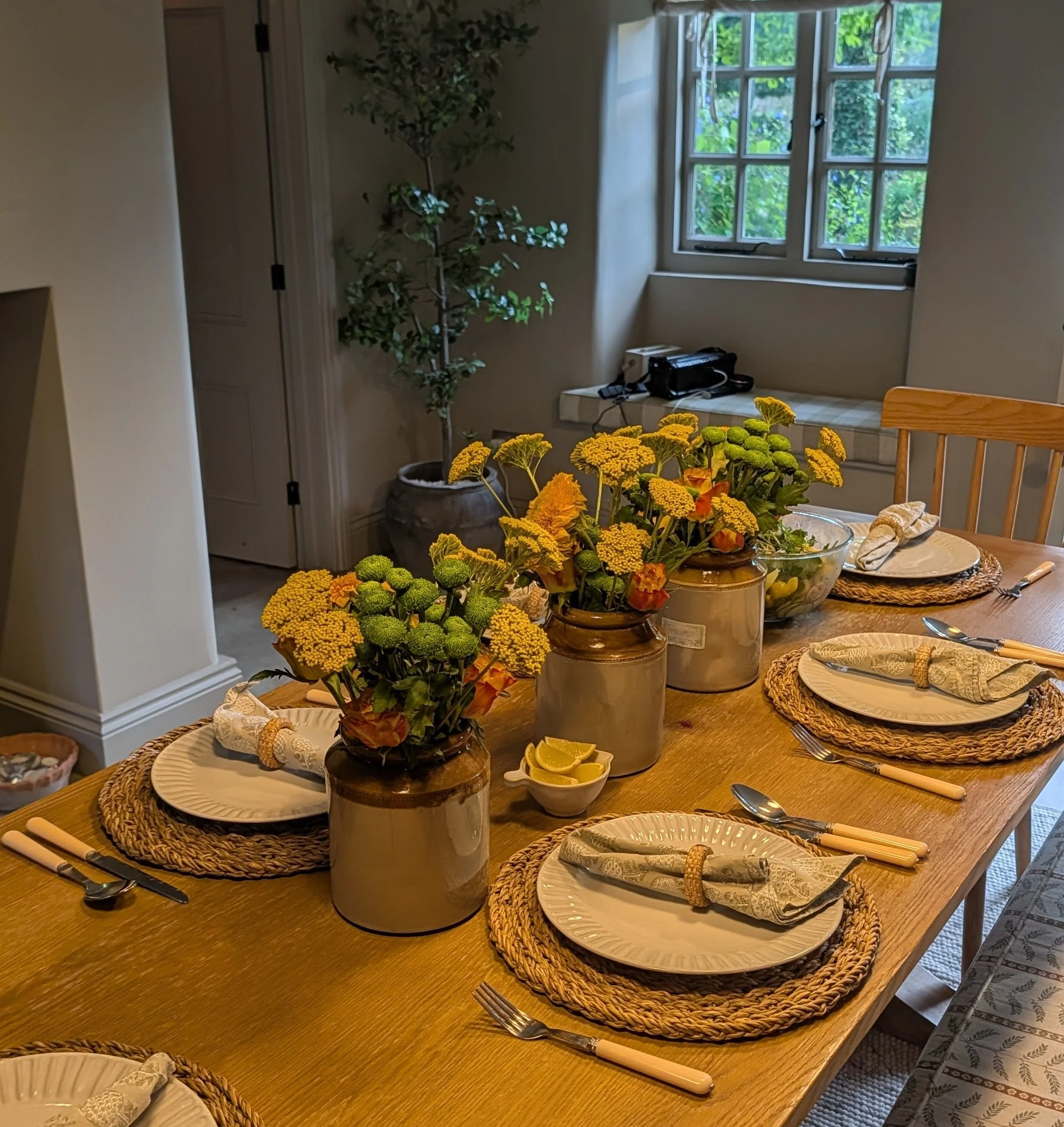 A dining table set with plates, napkins, and utensils, with a centrepiece of yellow and orange flowers in glass vases. There is a window with greenery outside in the background, and additional decorative elements and furniture in the room.