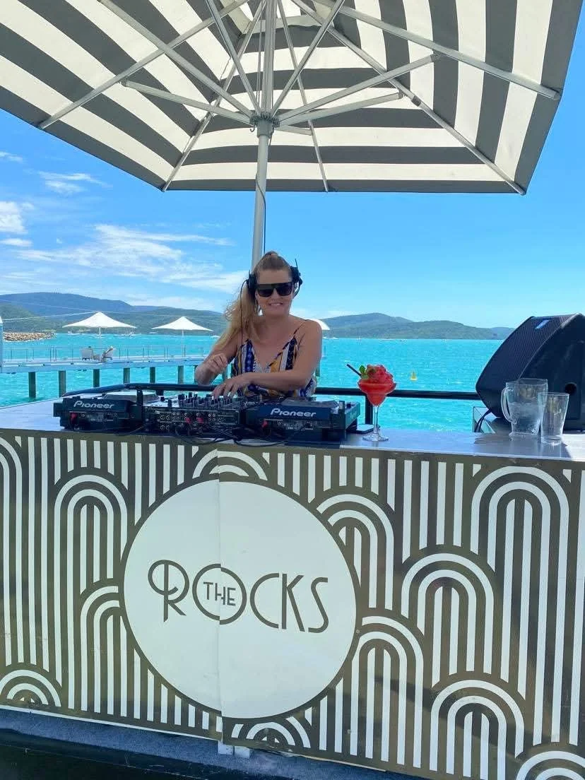 A DJ wearing sunglasses and headphones, standing behind a DJ booth with the logo "The Rocks", under a large striped umbrella, with a scenic ocean view and mountains in the background, on a sunny day.